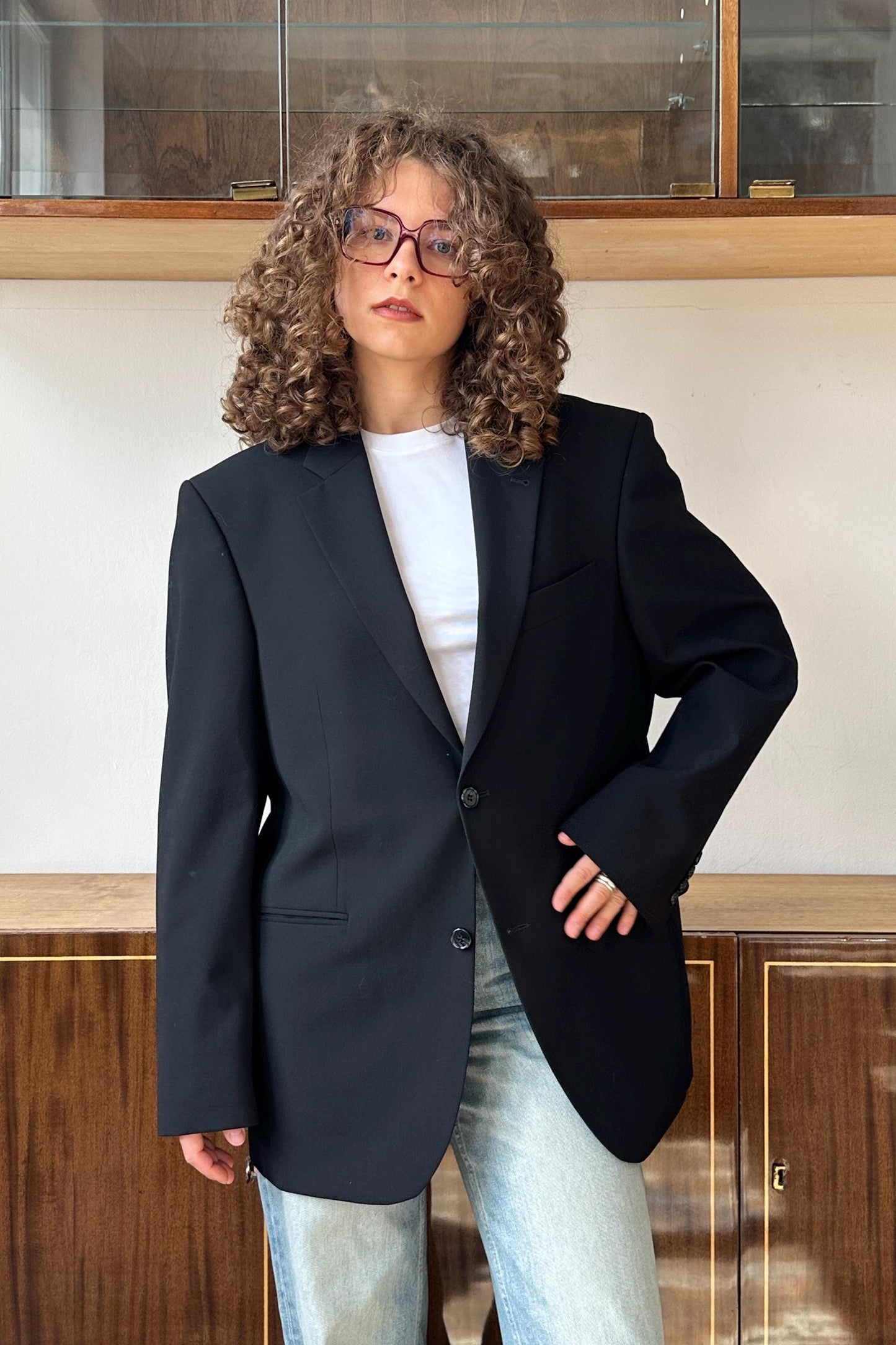 A woman with curly hair wearing a black blazer and glasses stands in front of a wooden cabinet.
