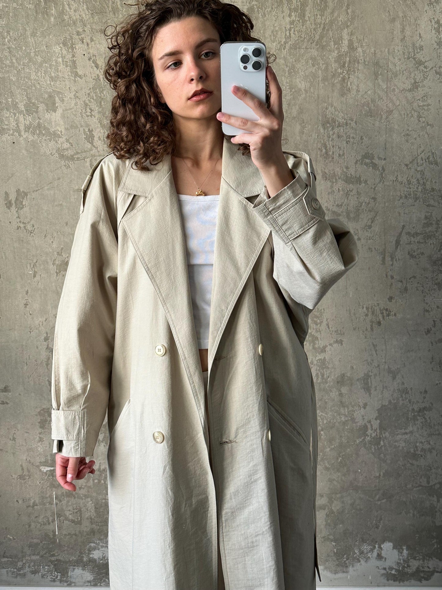 A woman with curly hair is taking a selfie with a white iPhone, wearing a beige trench coat.
