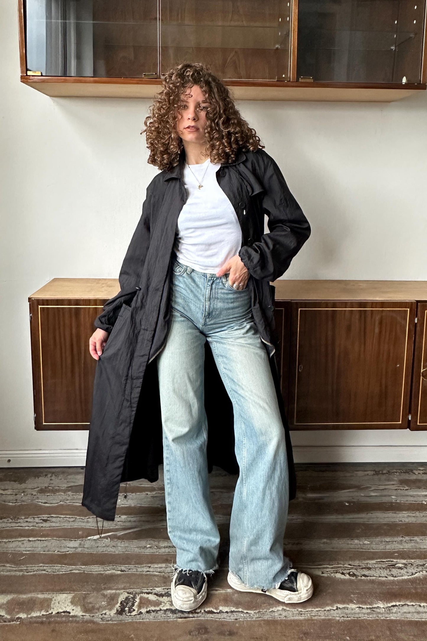 A woman with curly hair wearing a black coat, white shirt, and blue jeans stands in a room with wooden cabinets.