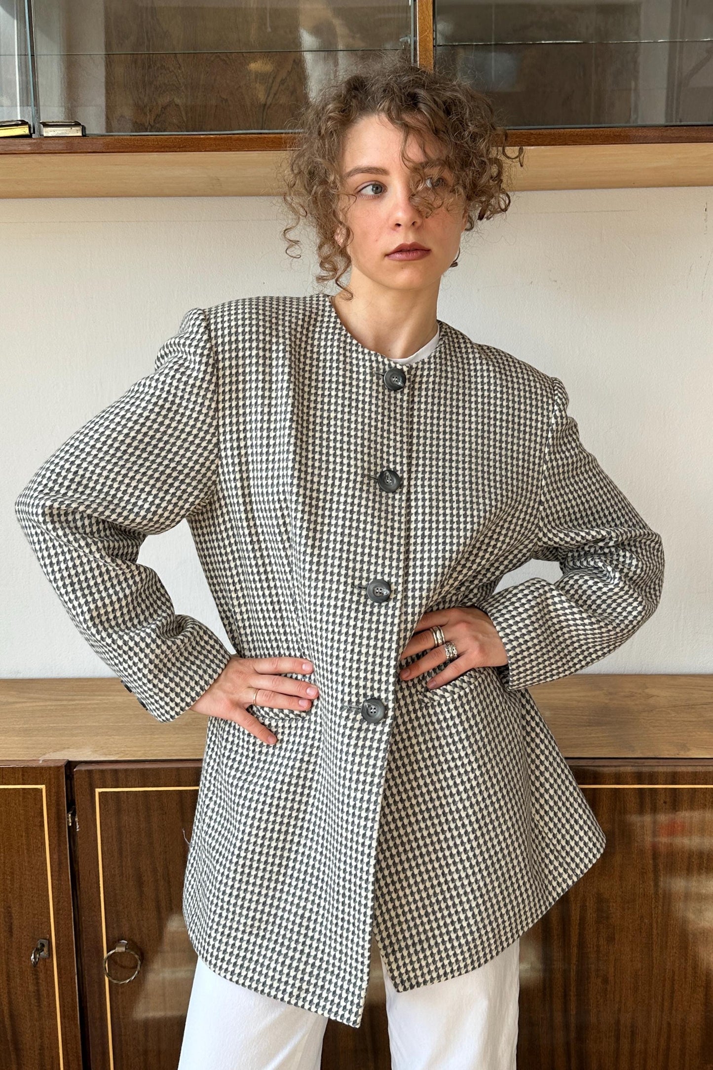 A woman with curly hair wearing a black and white checkered jacket stands with her hands on her hips, looking directly at the camera.