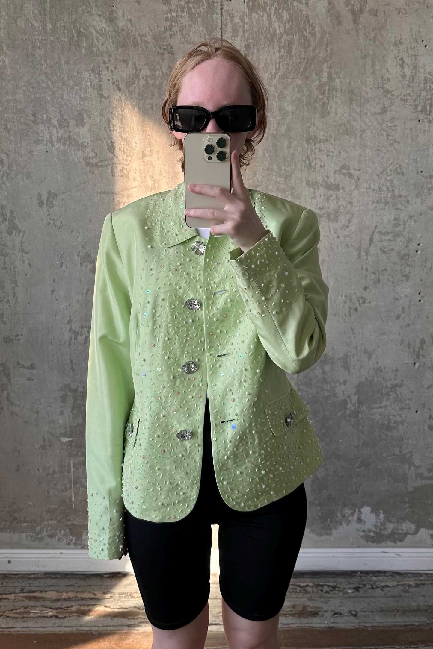 A person wearing a green jacket with sparkly details and sunglasses is taking a selfie with a smartphone.