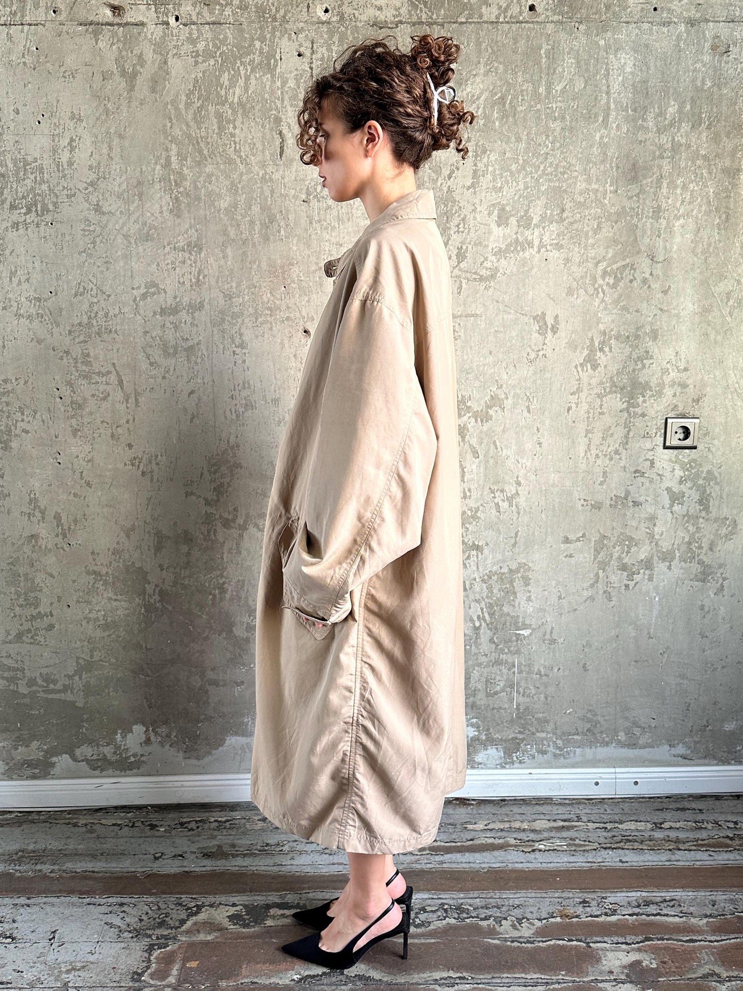 A woman wearing a beige trench coat stands in front of a concrete wall, with her hair styled in a bun.