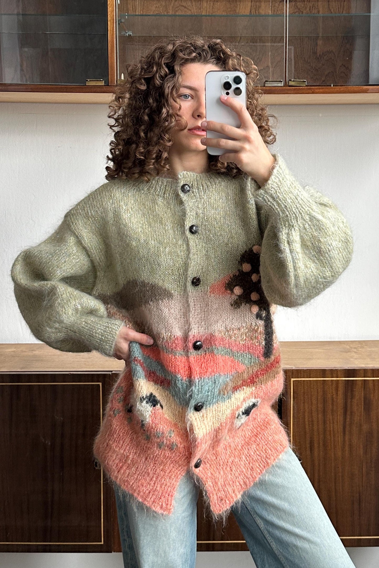 A woman with curly hair is taking a selfie with a smartphone, wearing a colorful, patterned cardigan.
