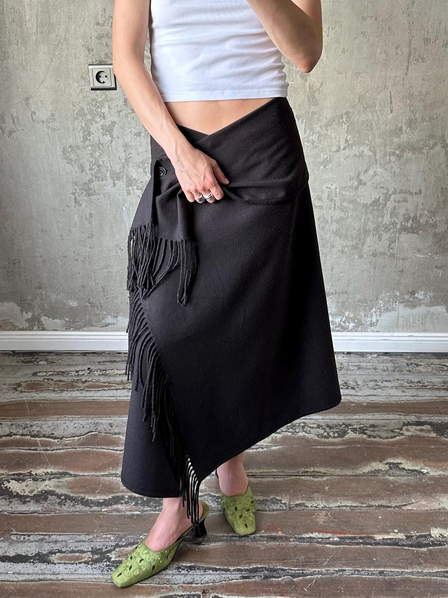 A person wearing a black skirt with fringe details and green shoes stands in front of a concrete wall.