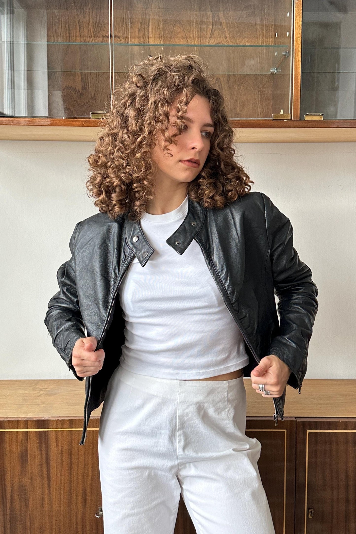 A woman with curly hair wearing a black leather jacket and white pants stands in front of a wooden cabinet.