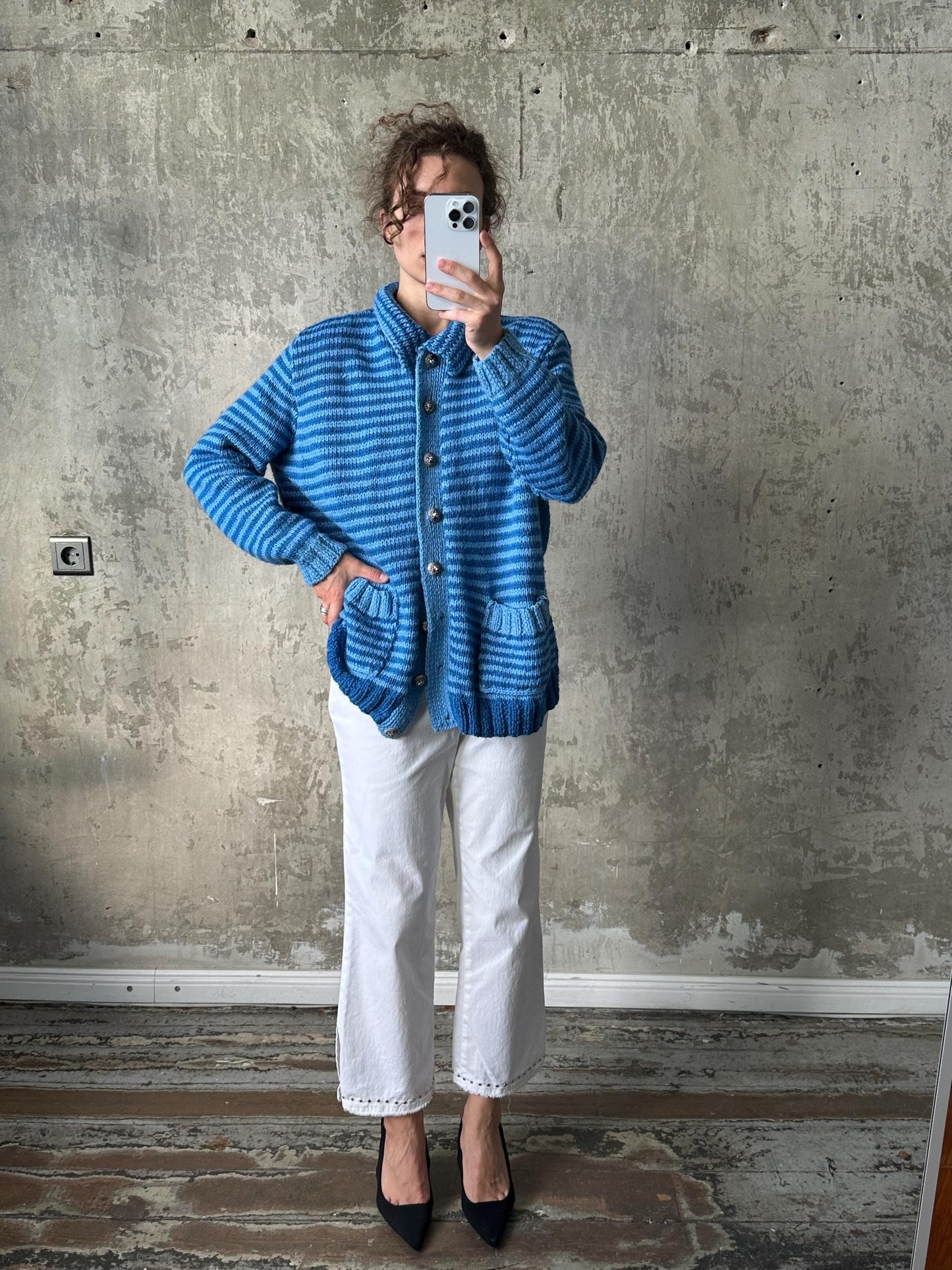 A person is taking a selfie with a blue striped cardigan and white pants, standing in front of a concrete wall.