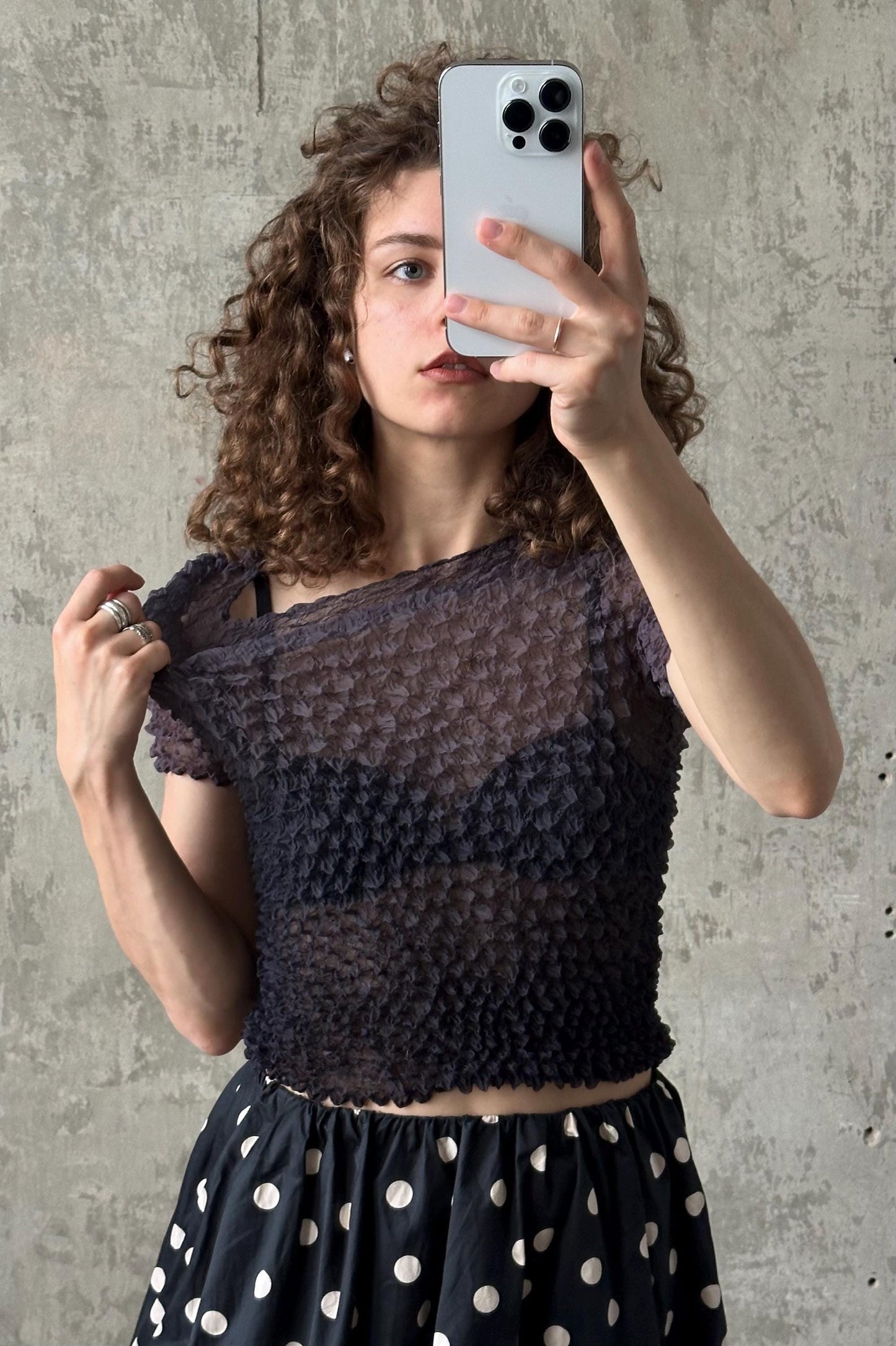 A young woman with curly hair is taking a selfie with a smartphone, wearing a black and white polka dot skirt and a black top with a ruffled texture.