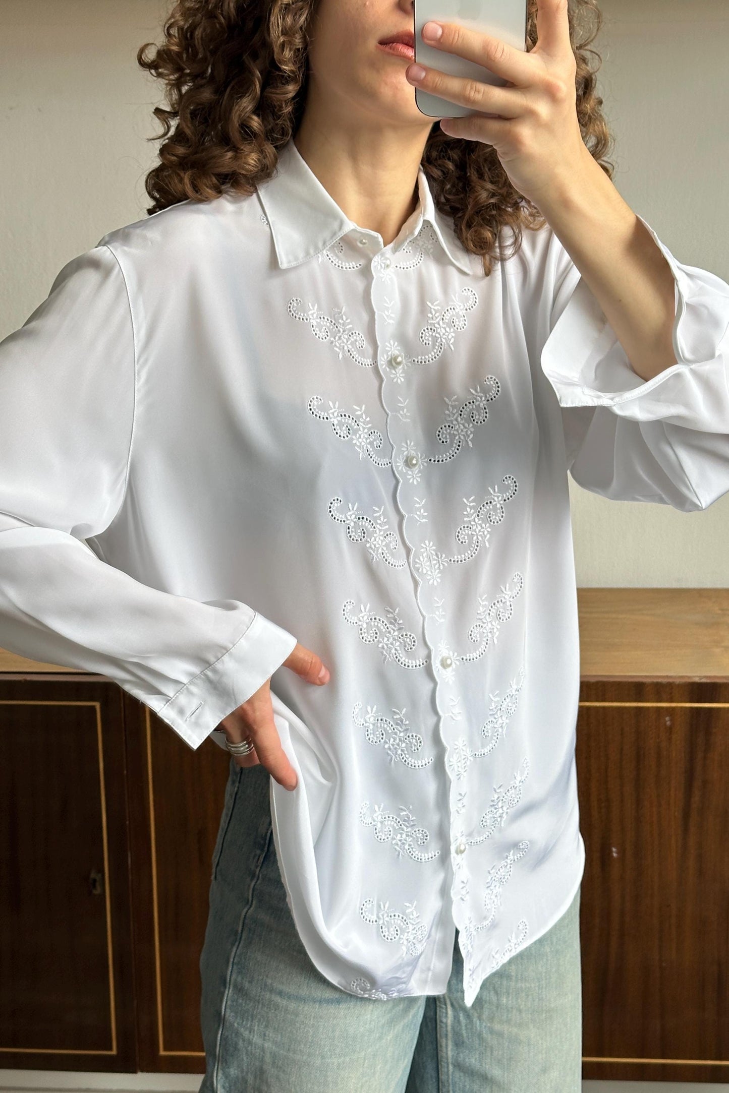 A woman with curly hair is taking a selfie with a white blouse featuring a floral pattern. She is holding a phone up to her face, capturing the image.