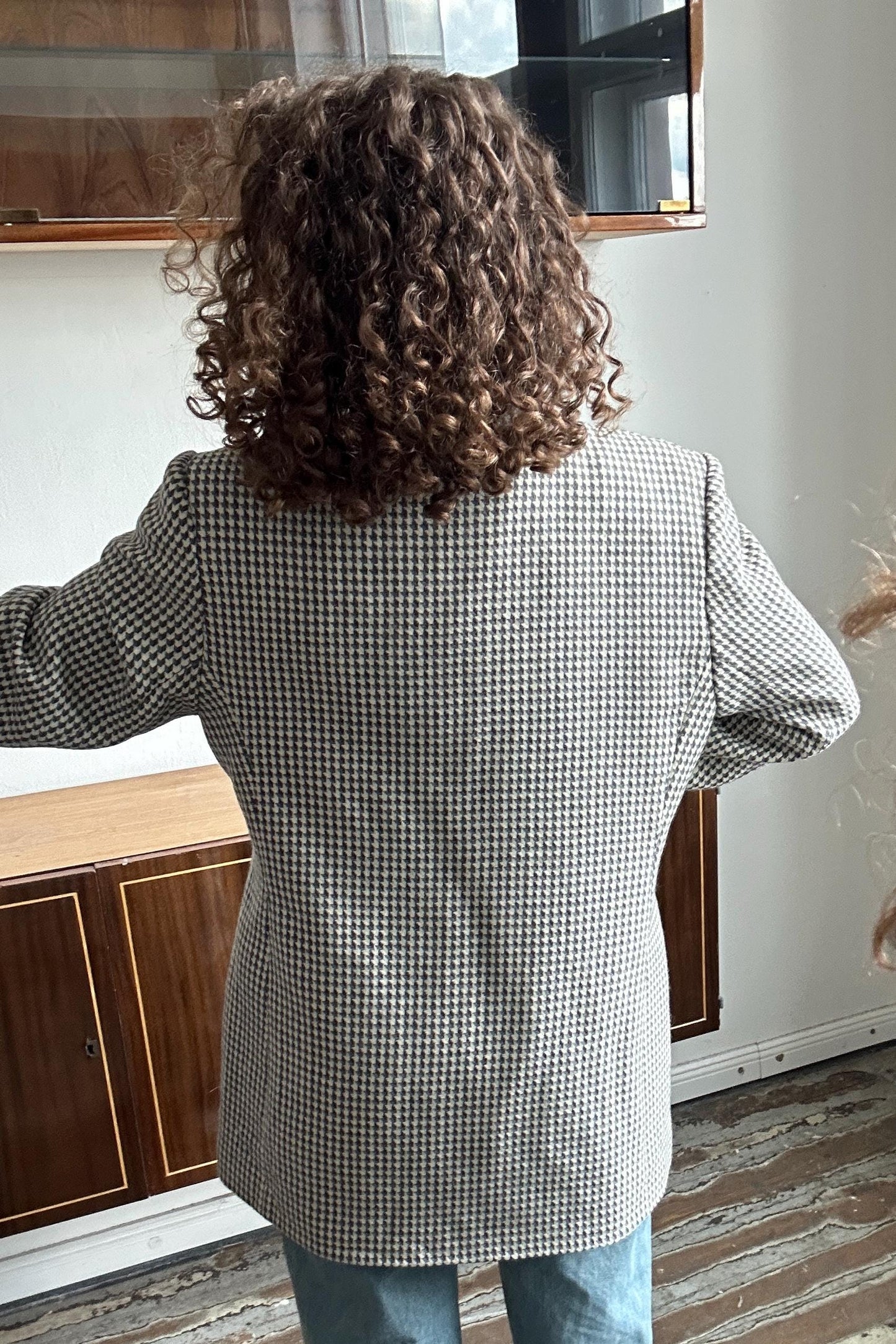 a person with curly hair wearing a black and white checkered jacket, standing in a room with a wooden cabinet and a mirror in the background.