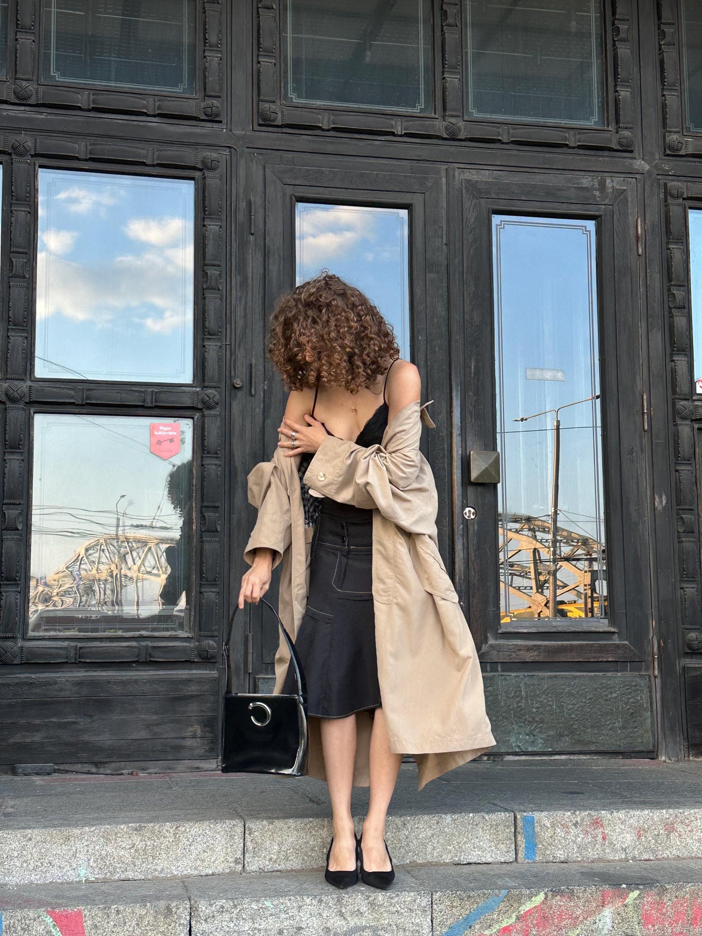 A woman with curly hair stands on a set of stairs, wearing a beige coat and holding a black purse.