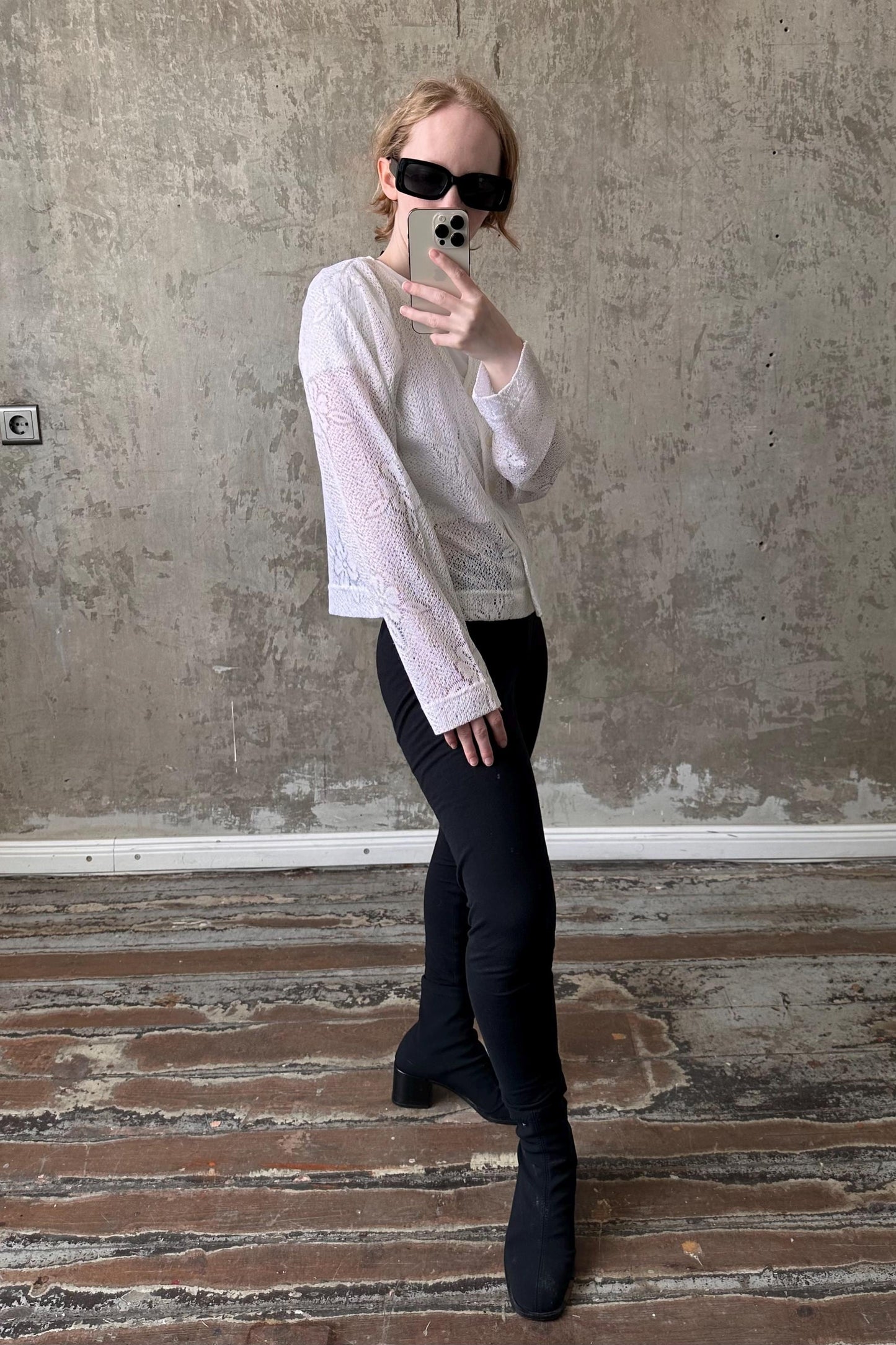 A person is taking a selfie with a smartphone, wearing a white lace top, black pants, and black boots, standing in front of a concrete wall.