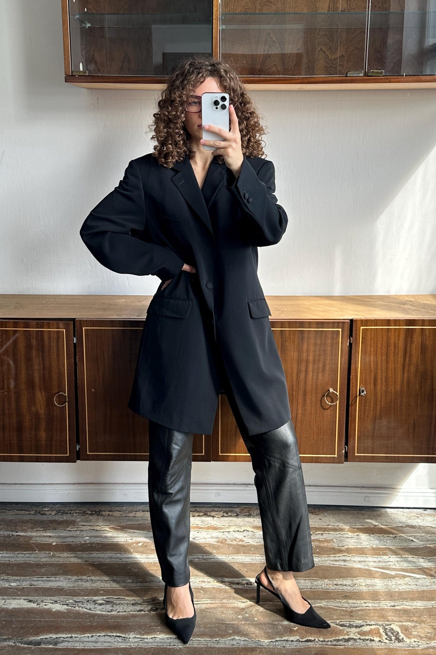 A woman with curly hair is taking a selfie with a smartphone, wearing a black blazer and leather pants, standing in front of a wooden cabinet.