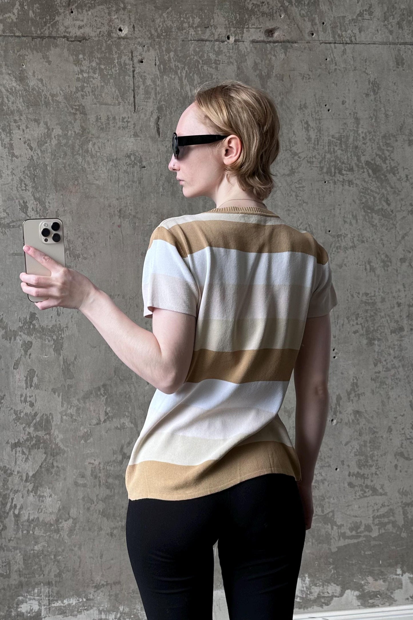 A woman wearing sunglasses and a striped shirt is holding a smartphone and taking a selfie in front of a concrete wall.