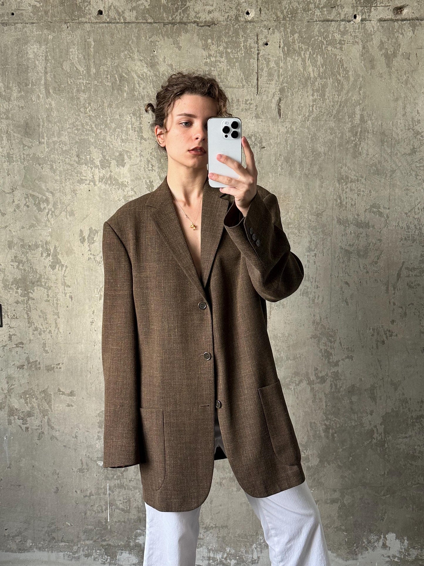 A person is taking a selfie with a white phone, wearing a brown blazer and white pants against a concrete wall background.