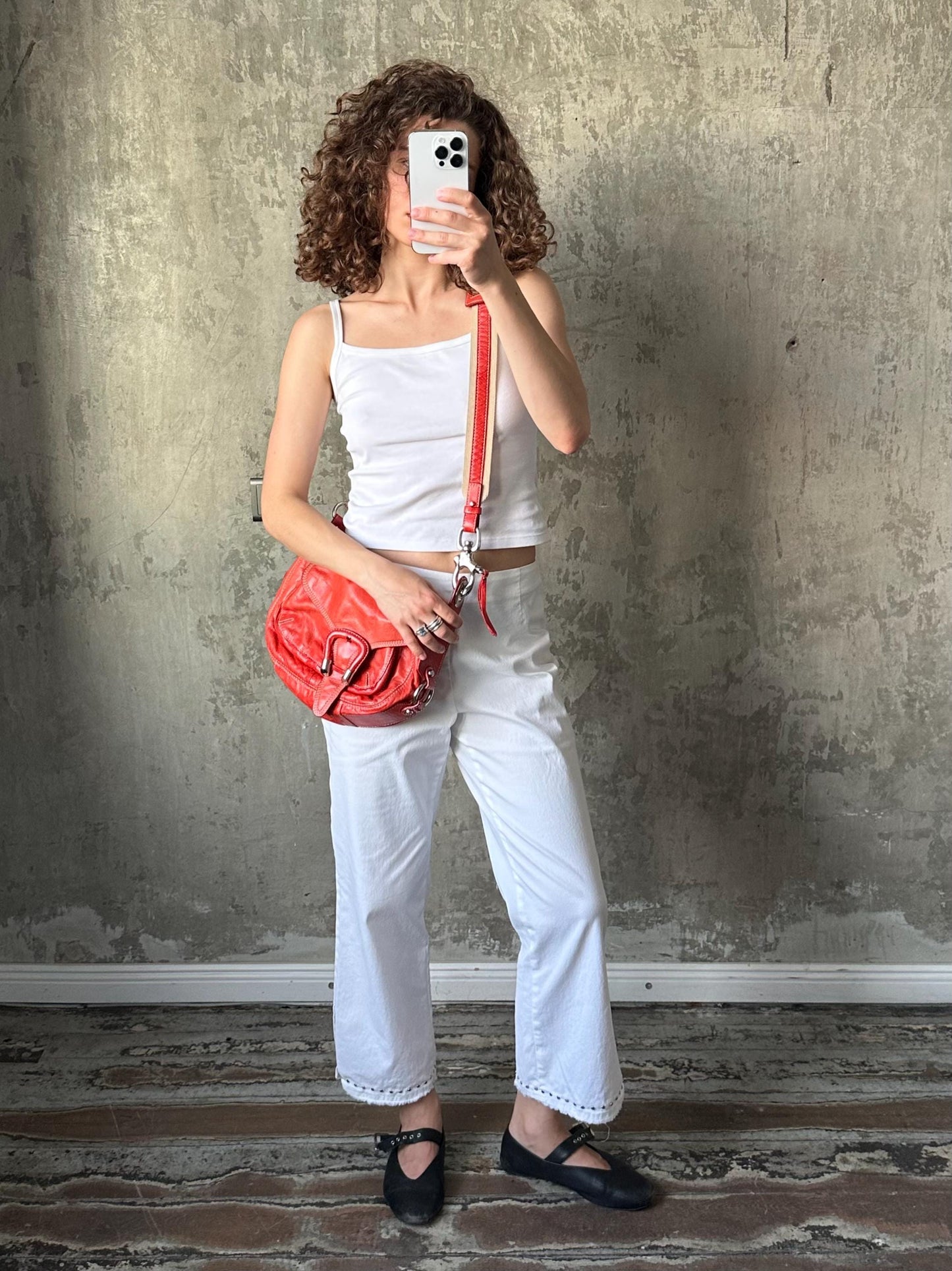 A woman with curly hair is taking a selfie with a white iPhone, holding a red purse, and wearing white pants and black shoes.