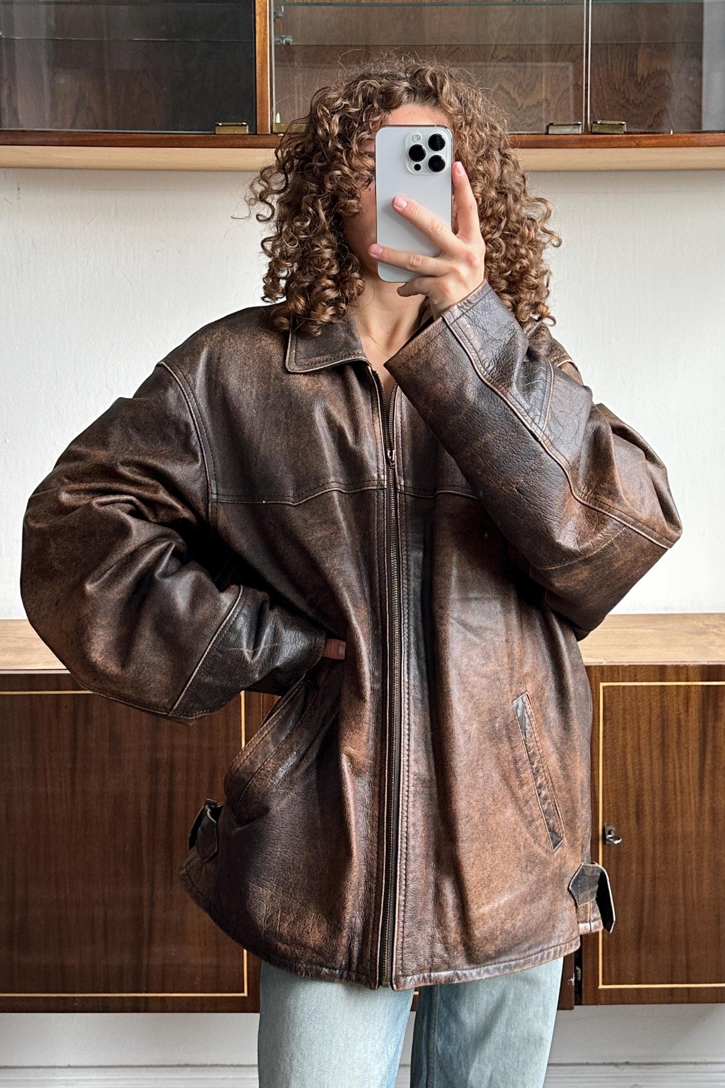 A person with curly hair is taking a selfie with a smartphone, wearing a brown leather jacket.