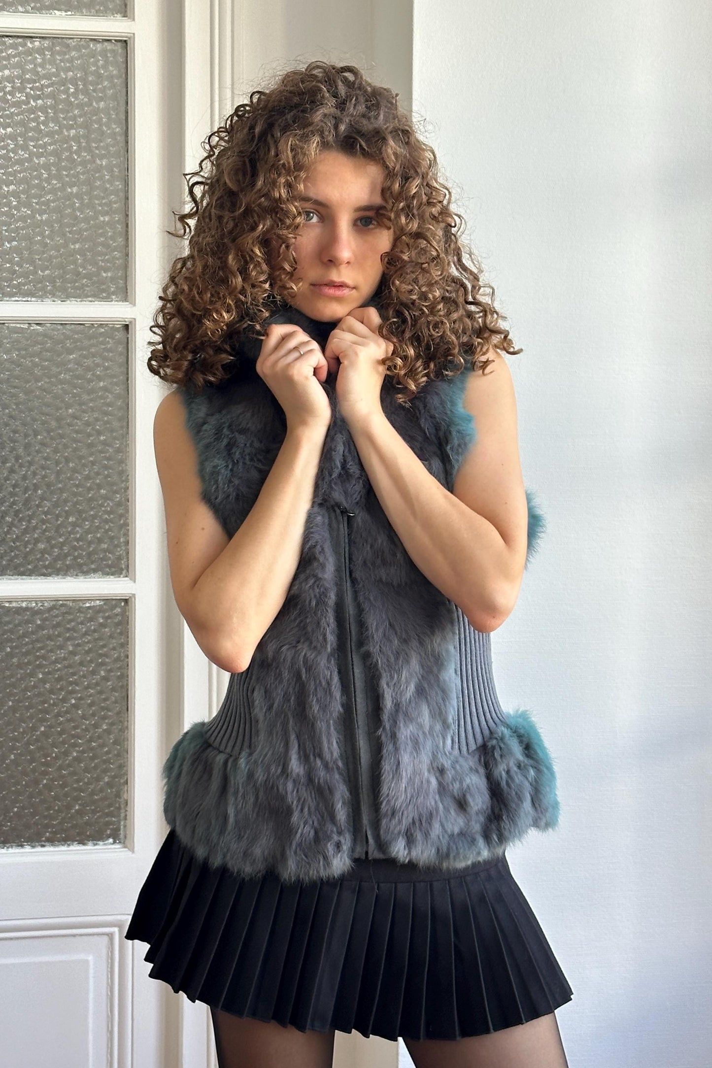 A young woman with curly hair stands in front of a white door, wearing a gray fur vest and a black pleated skirt.