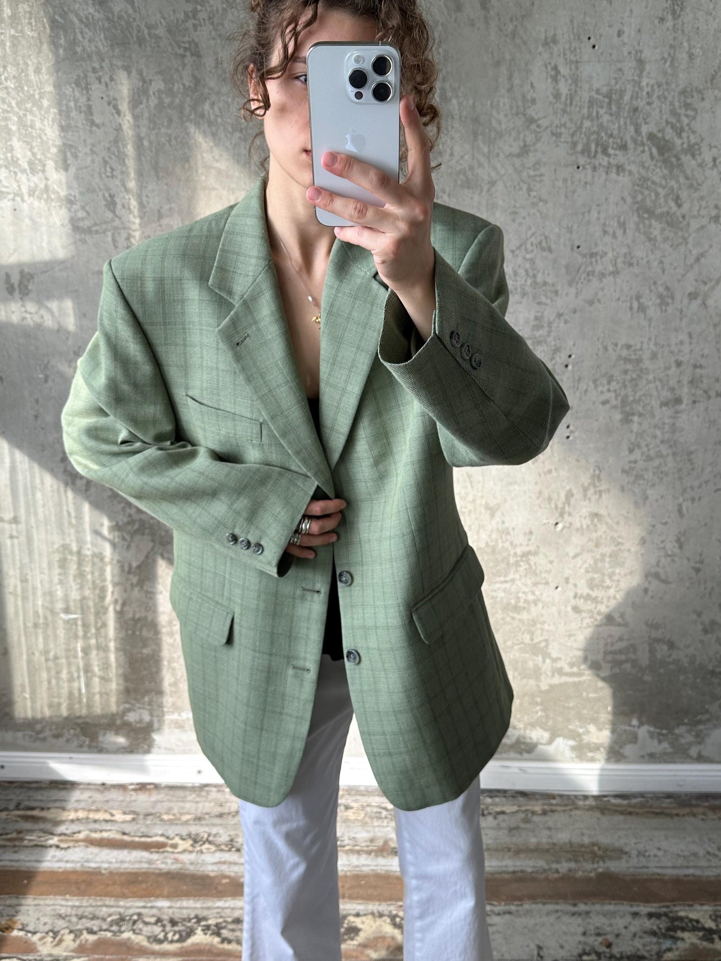 A person is taking a selfie with a smartphone, wearing a green plaid blazer and white pants.