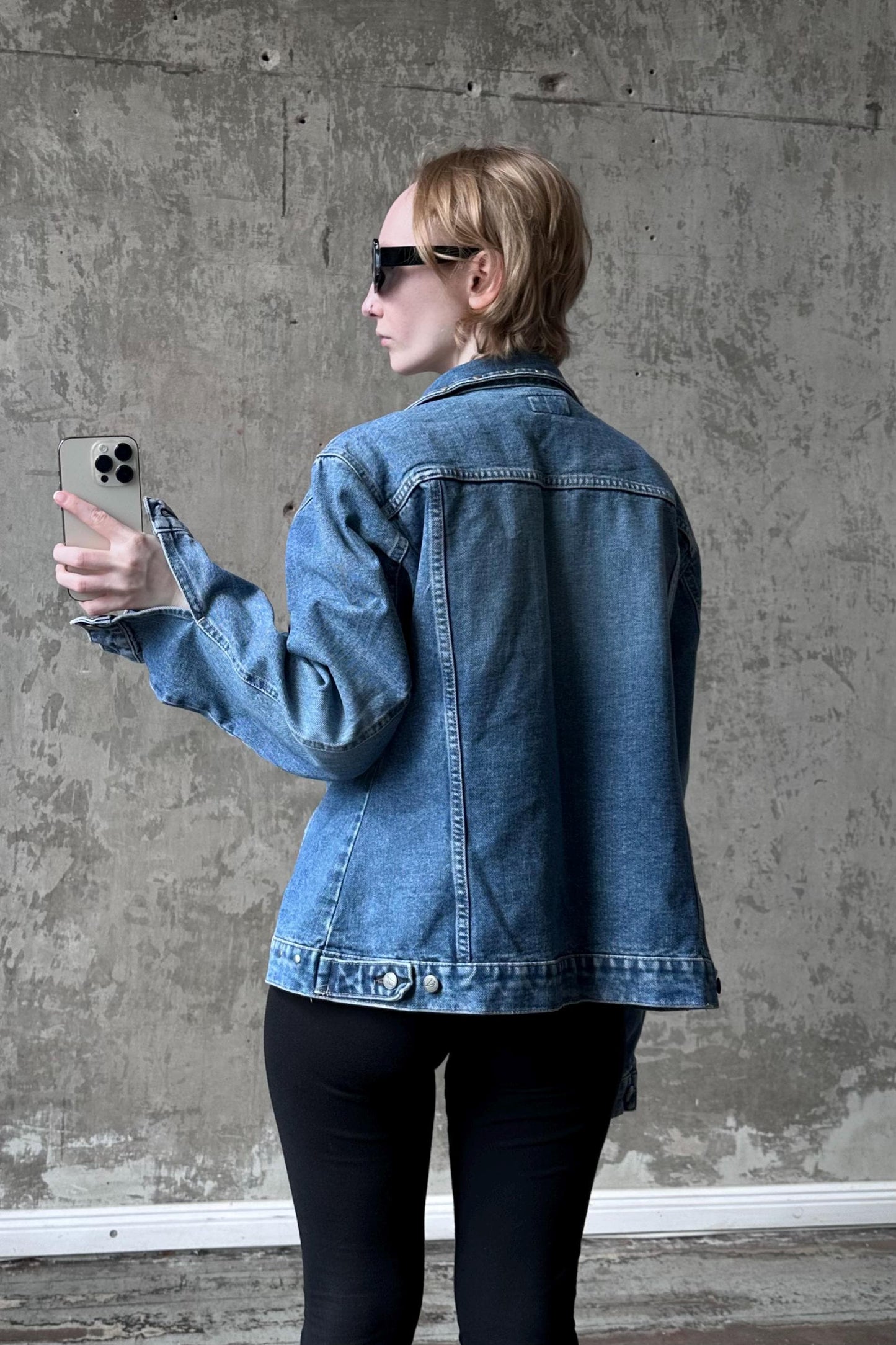 A woman wearing a denim jacket and sunglasses is holding a smartphone and looking at the screen.