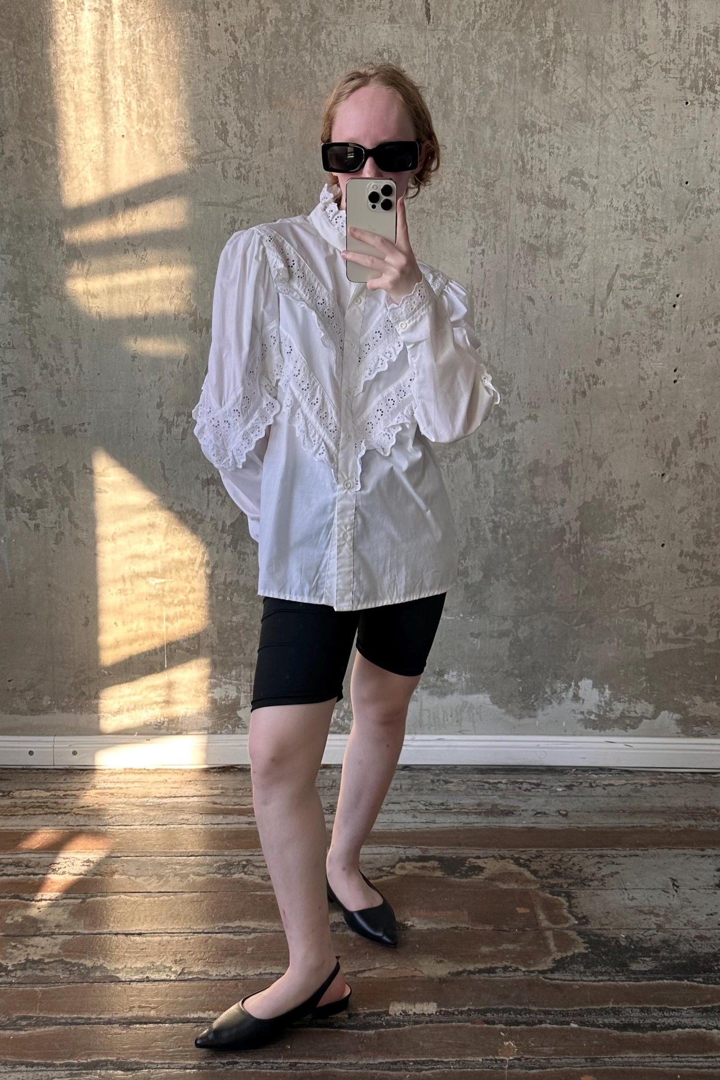 A person is taking a selfie with a smartphone, wearing a white blouse, black shorts, and black shoes, standing in front of a concrete wall.