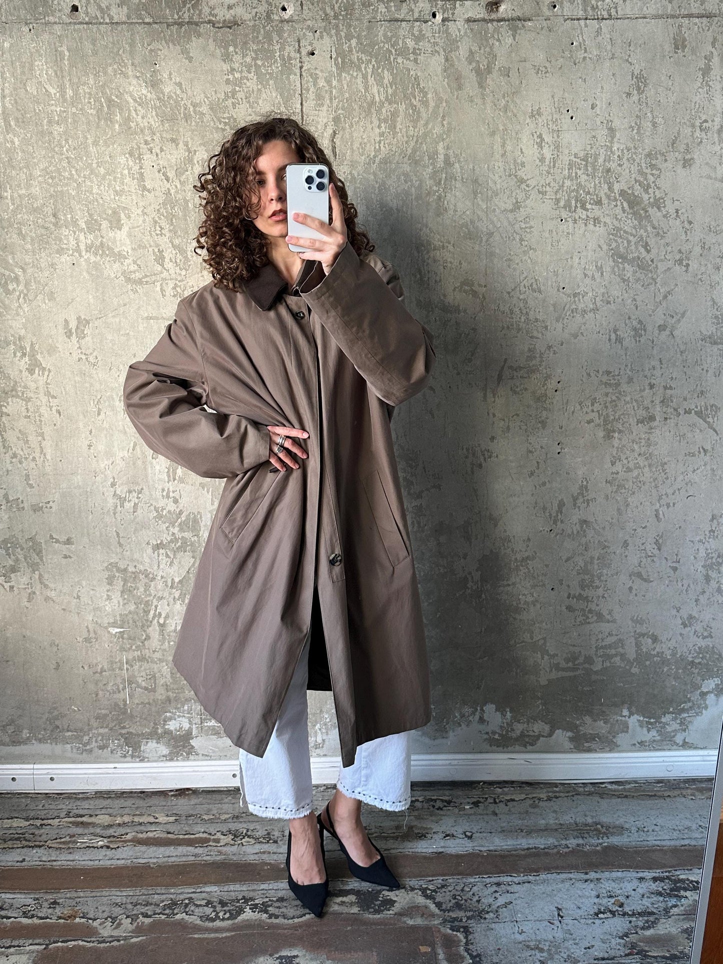 A woman in a brown coat is taking a selfie with a white phone against a concrete wall.