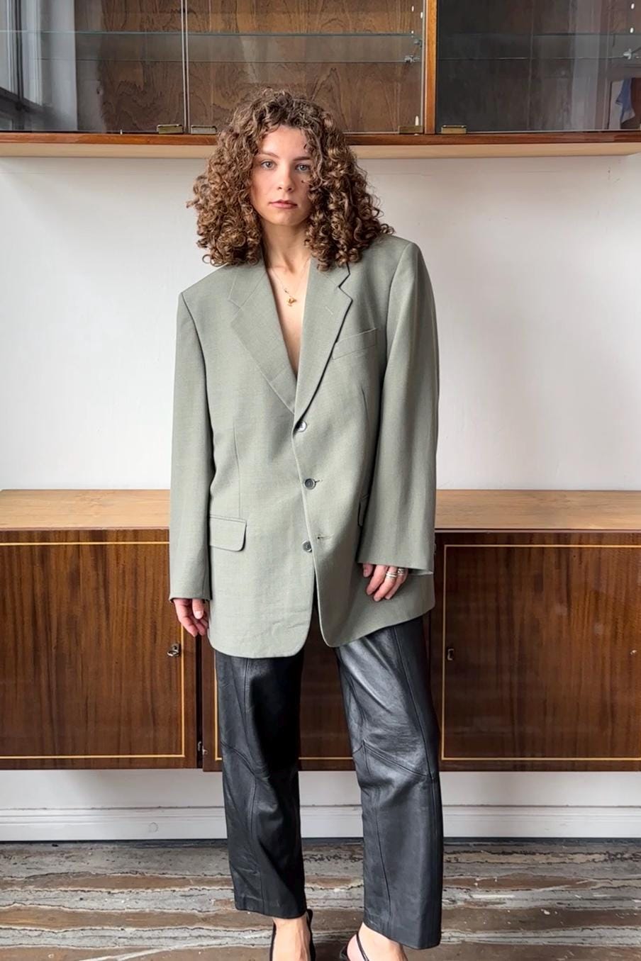 A woman with curly hair wearing a gray blazer and black pants stands in front of a wooden cabinet.