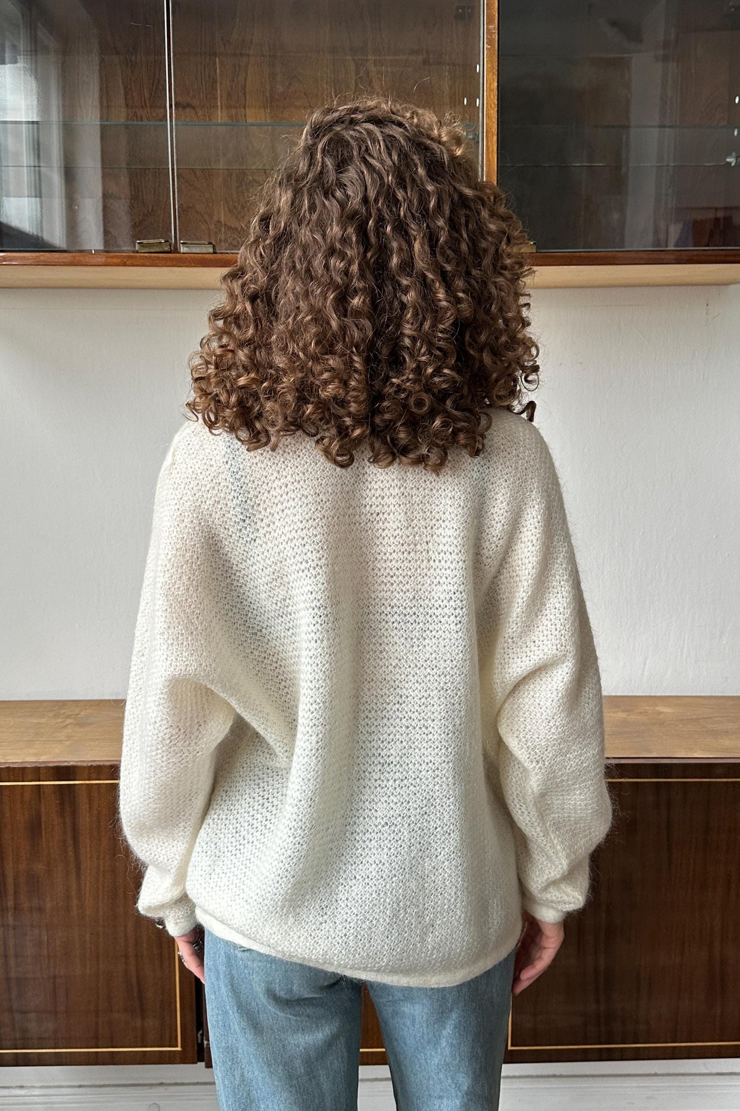 a person with curly hair wearing a white sweater, standing in front of a glass display case.
