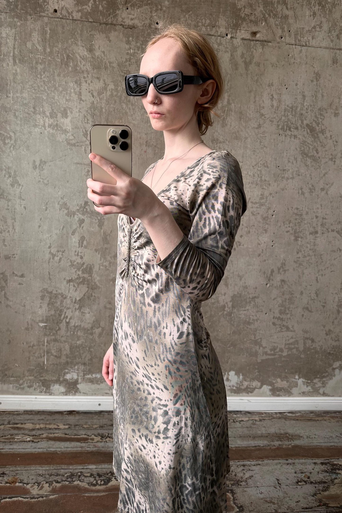 A woman wearing a leopard print dress and sunglasses is taking a selfie with her iPhone against a concrete wall.