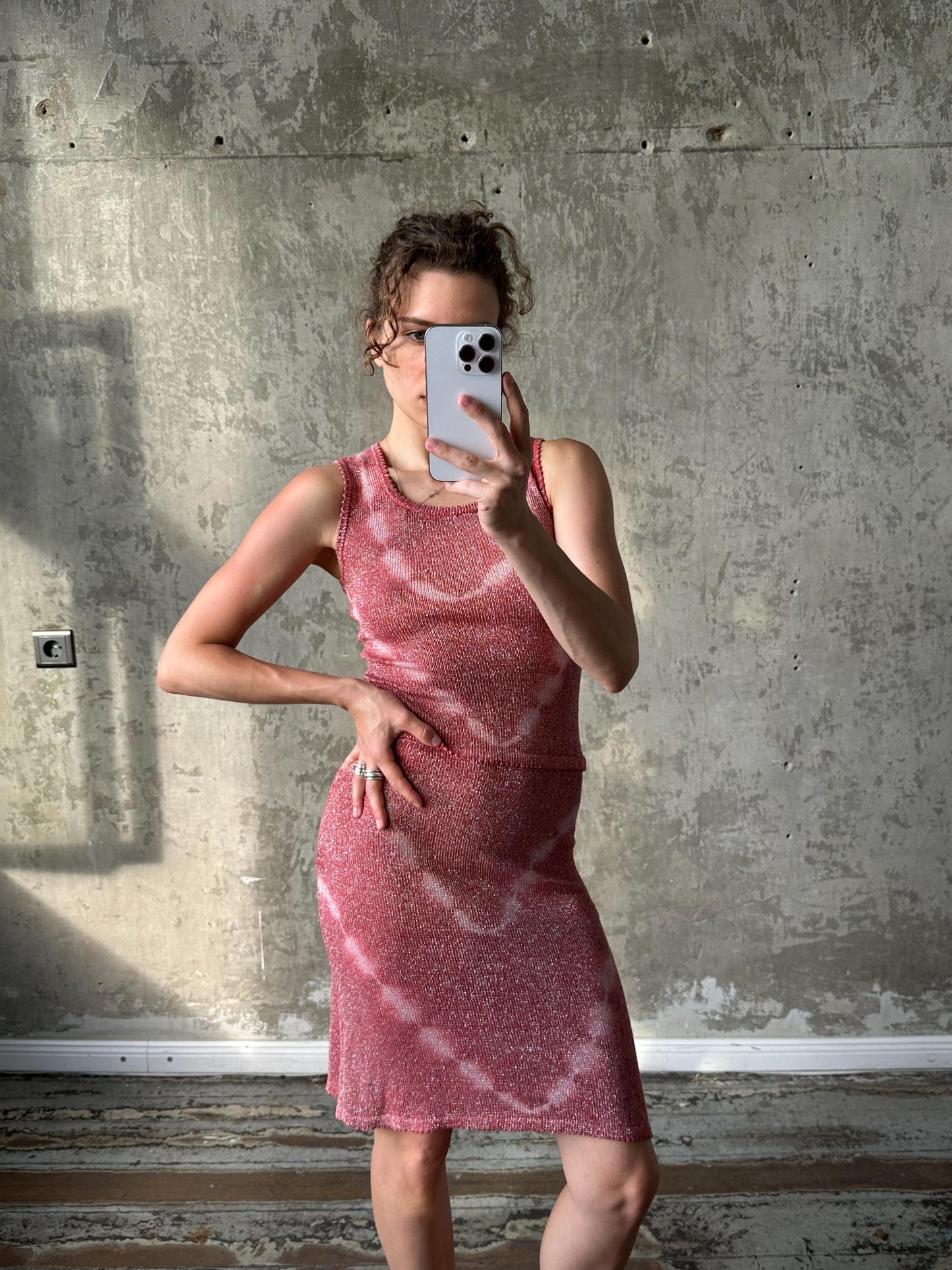 A woman in a pink dress is taking a selfie with a smartphone, standing in front of a concrete wall.