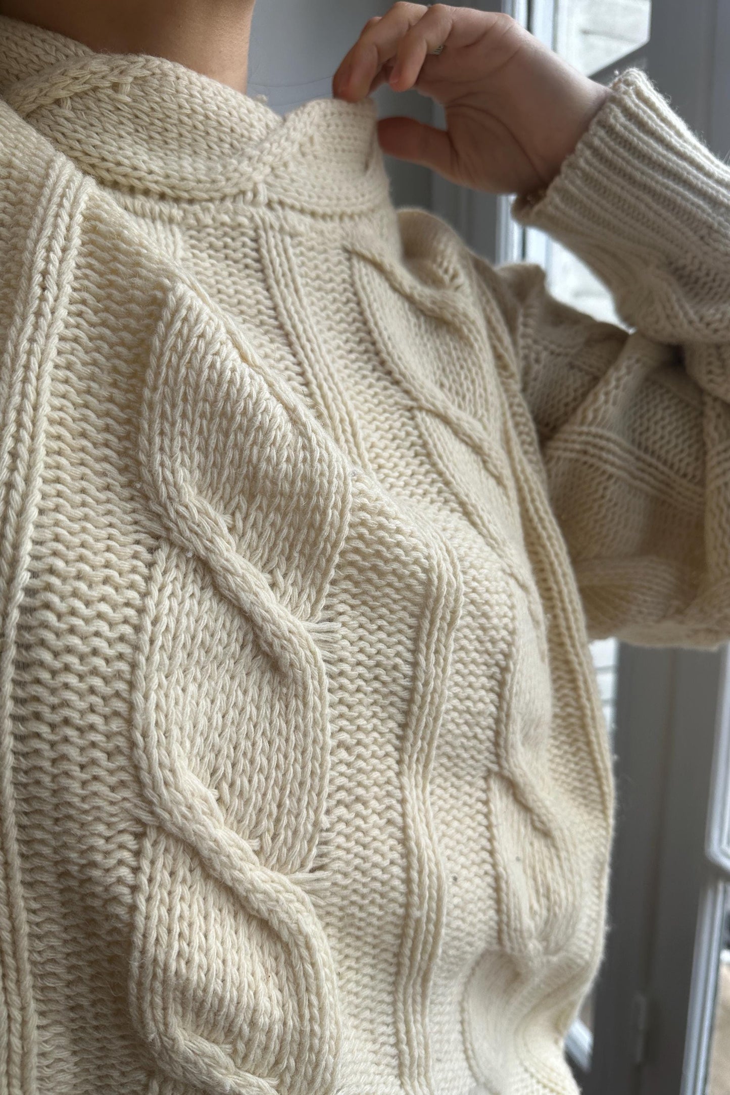 A close-up view of a person wearing a cream-colored knitted sweater, with their hand adjusting the collar.