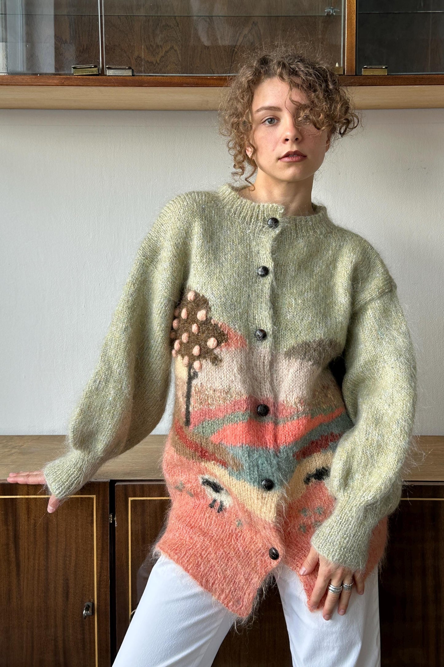 A woman wearing a light green cardigan with a colorful, abstract design stands in front of a wooden cabinet.
