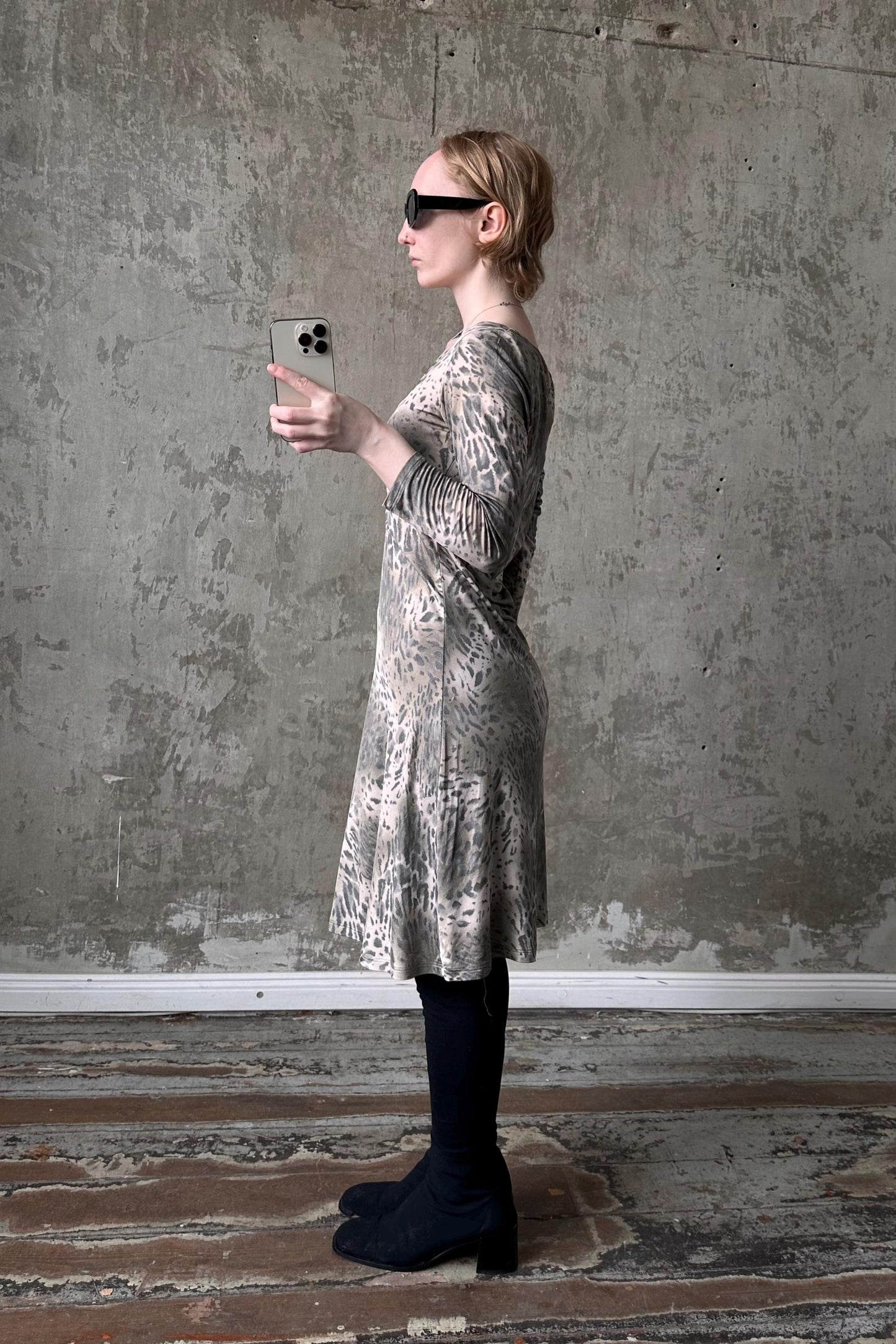 A woman in a leopard print dress stands in front of a concrete wall, holding a smartphone and looking at it.