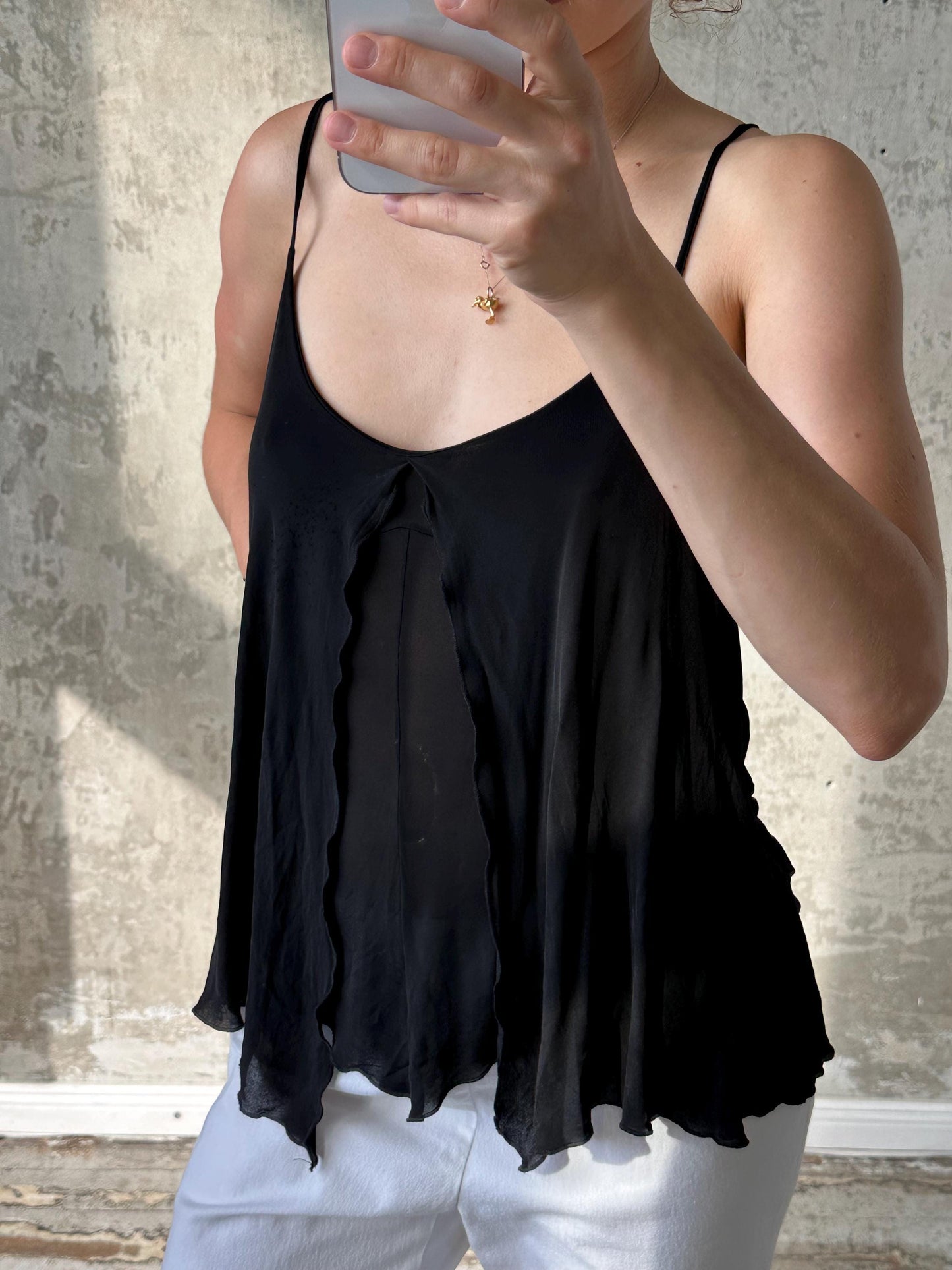 A person is taking a selfie with a black tank top and white pants, holding a smartphone in their hand.