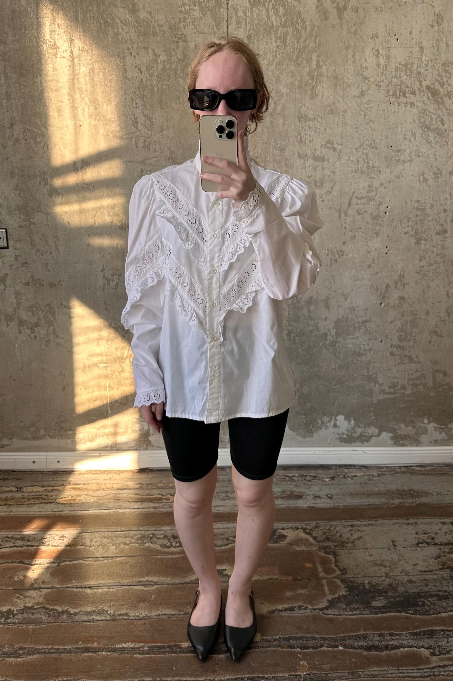 A person is taking a selfie with a white blouse and black shorts, standing in front of a concrete wall.