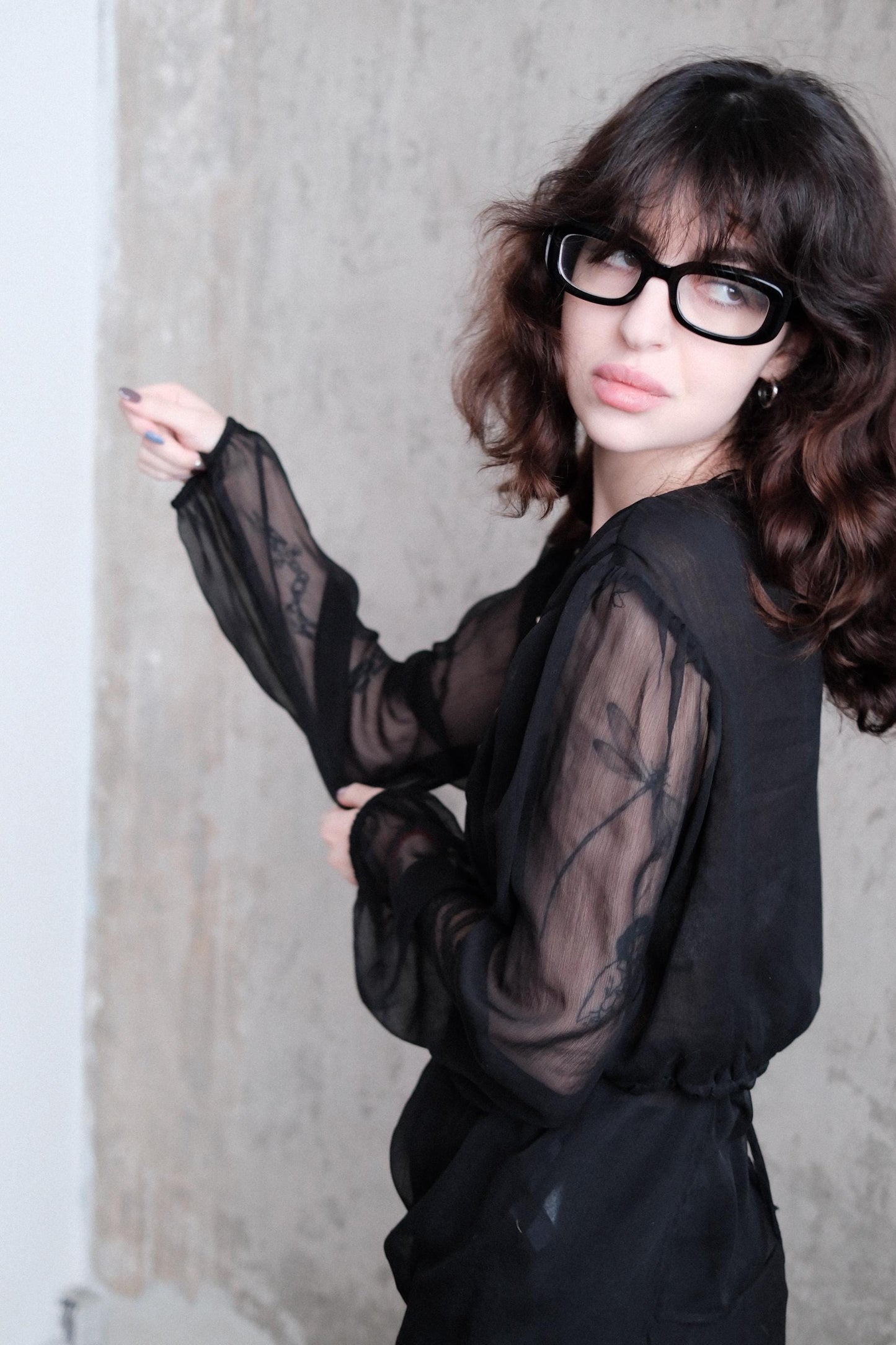 A woman with long, dark hair and glasses is lying on the ground, wearing a black dress with sheer sleeves.