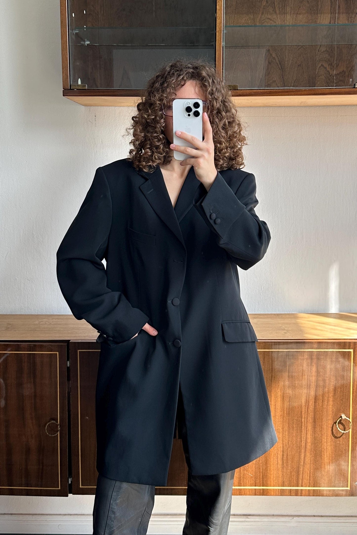 A person with curly hair is taking a selfie with a white phone, wearing a black blazer.