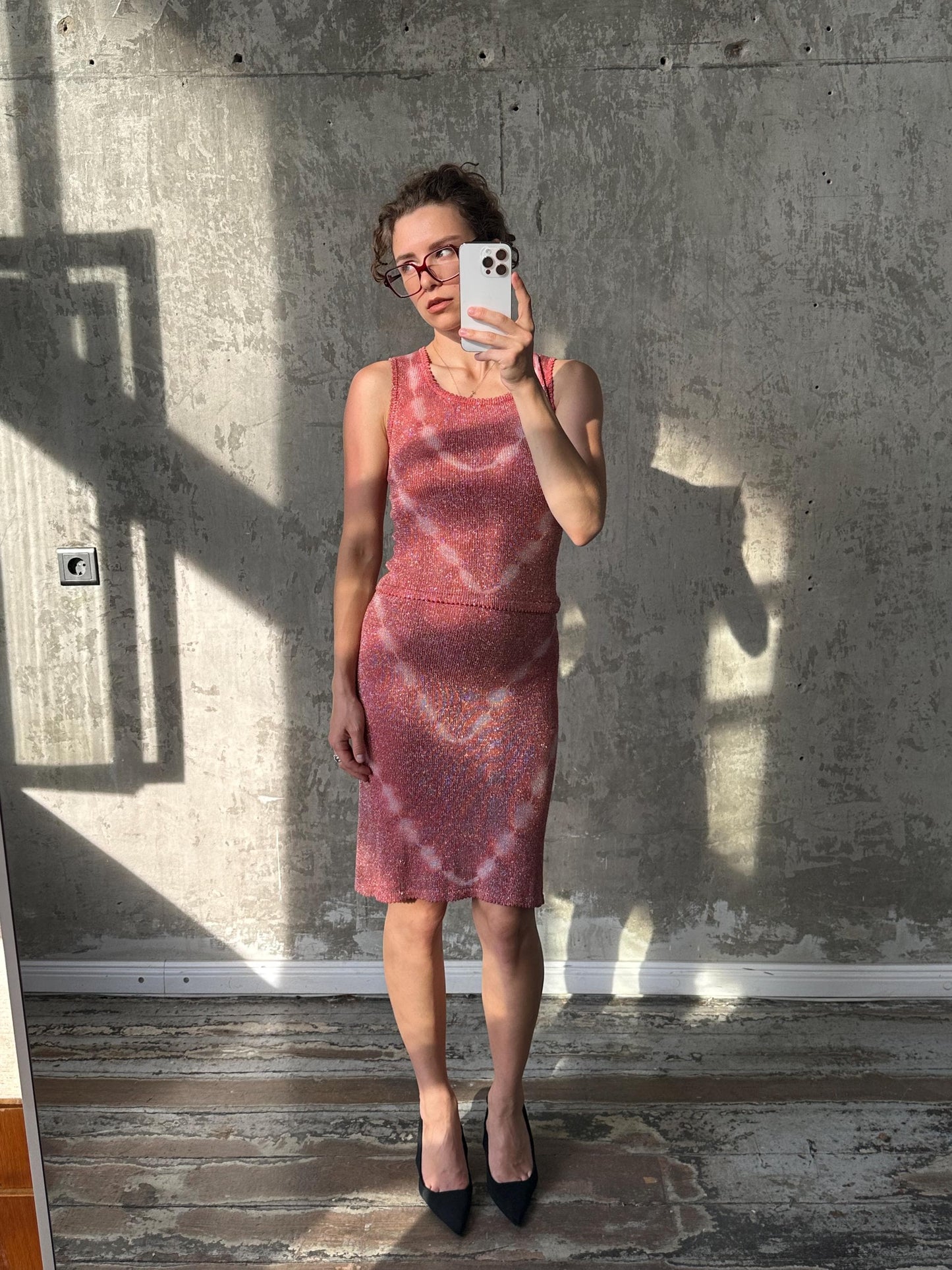 A woman in a pink dress is taking a selfie with a white phone, standing in front of a concrete wall.