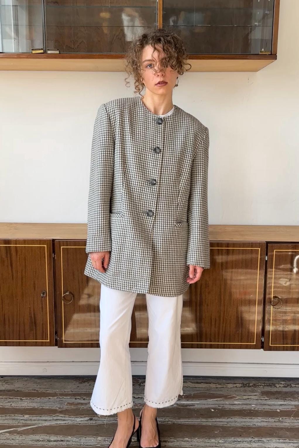 A woman wearing a checkered jacket and white pants stands in front of a wooden cabinet.