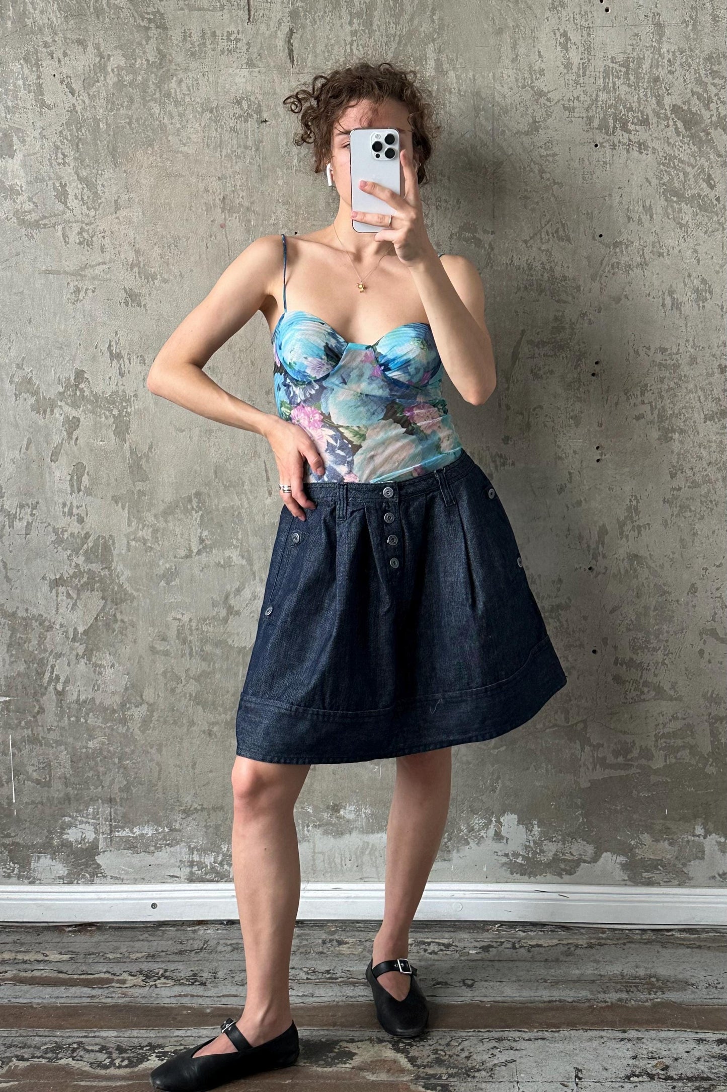 A woman is taking a selfie with her phone, wearing a blue and white patterned top and a denim skirt, standing in front of a concrete wall.