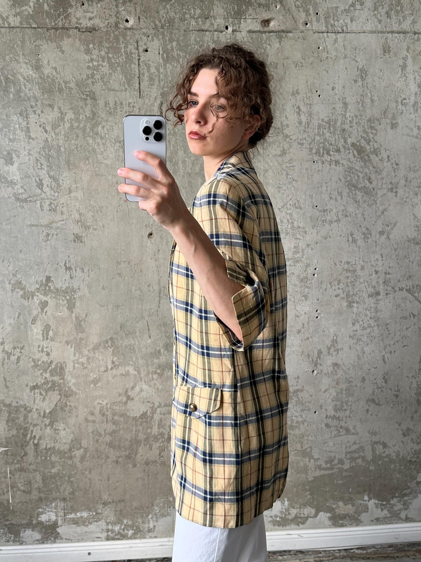 A person is taking a selfie with a smartphone, wearing a plaid jacket, against a concrete wall.