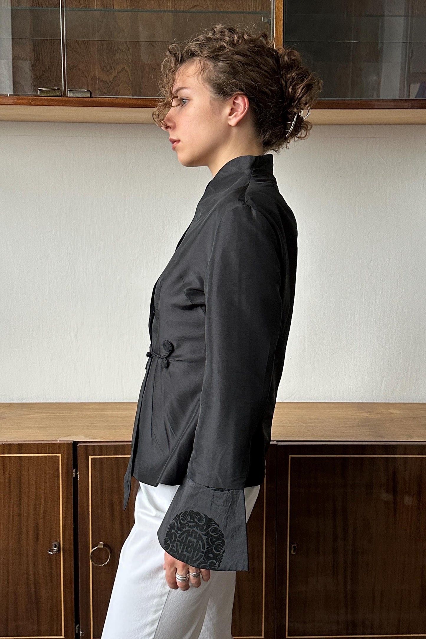 A woman wearing a black jacket with a black design on the sleeve stands in front of a wooden cabinet.
