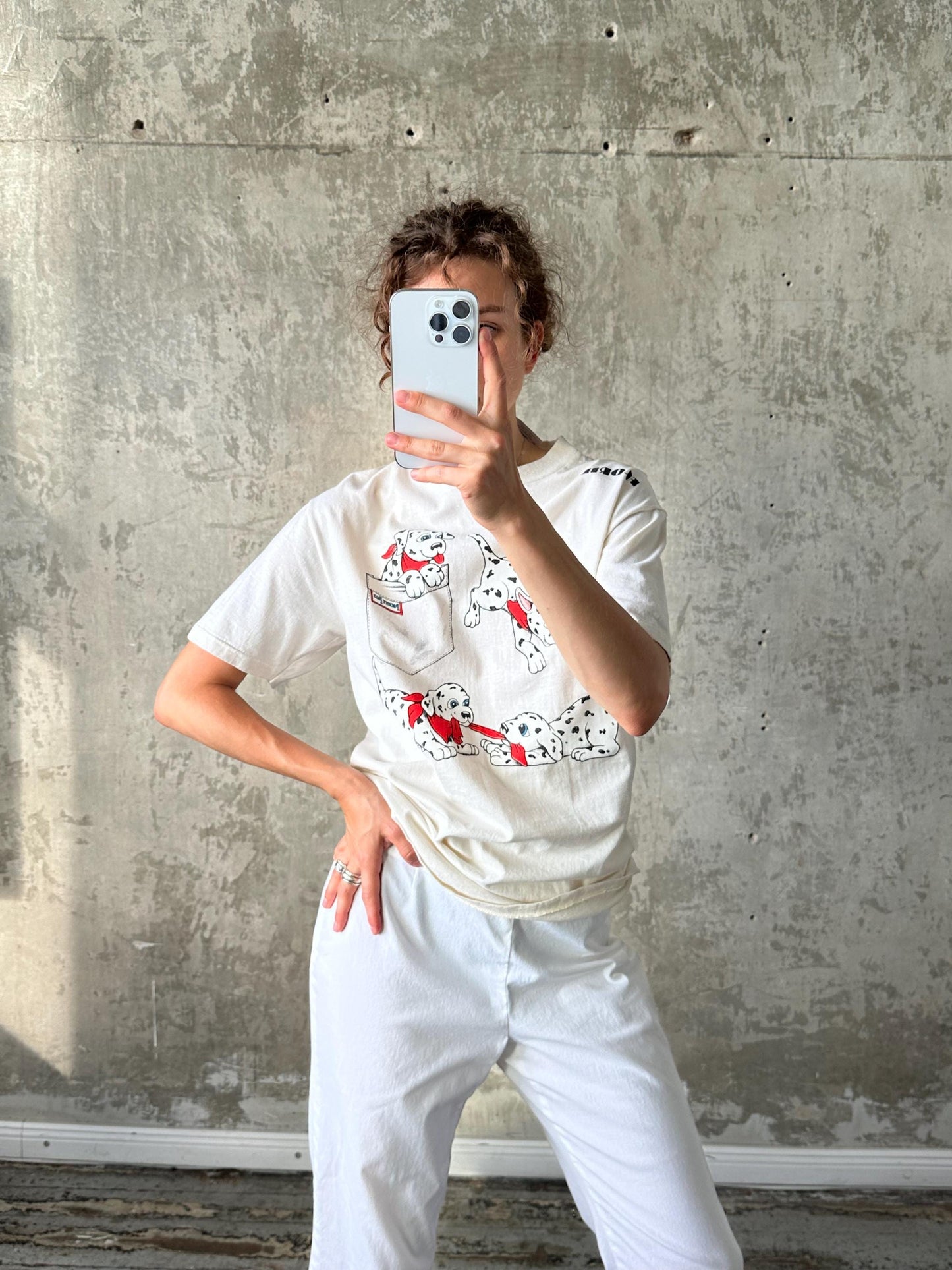 A person is taking a selfie with a smartphone, wearing a white t-shirt with a Dalmatian design and white pants.