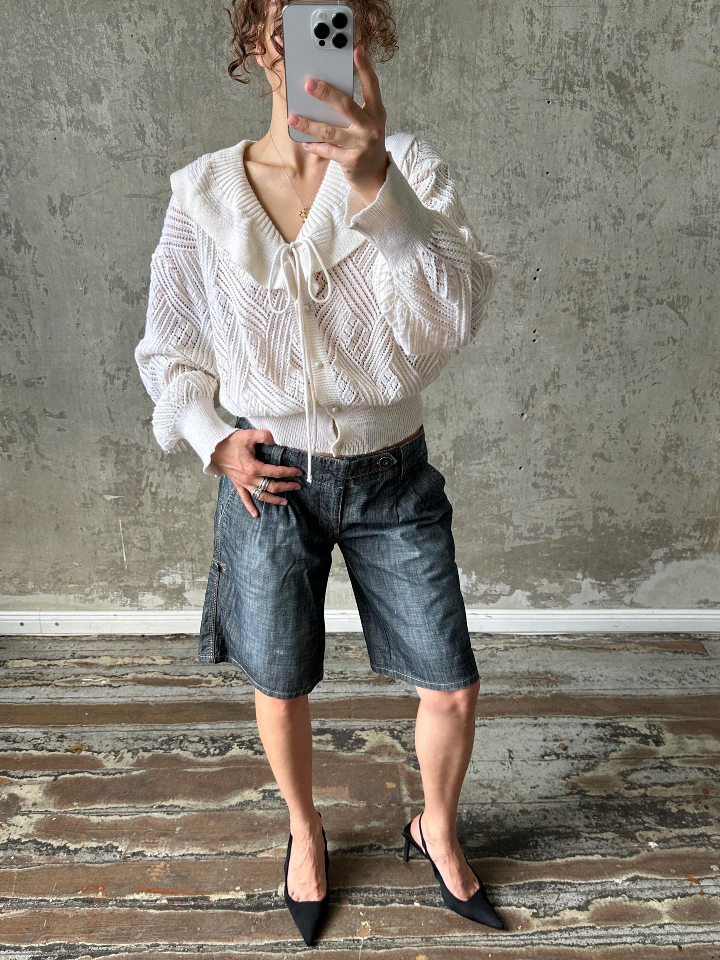 A person is taking a selfie with a smartphone, wearing a white sweater, blue jeans, and black heels.