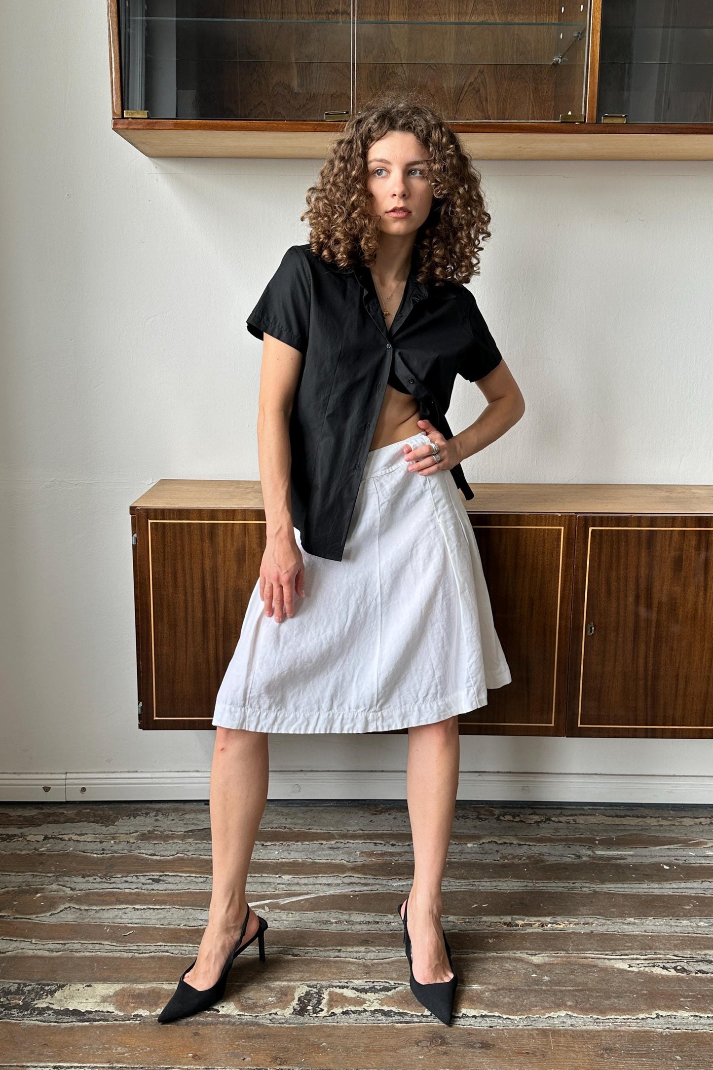 A woman with curly hair wearing a black shirt and white skirt stands in a room with wooden cabinets.