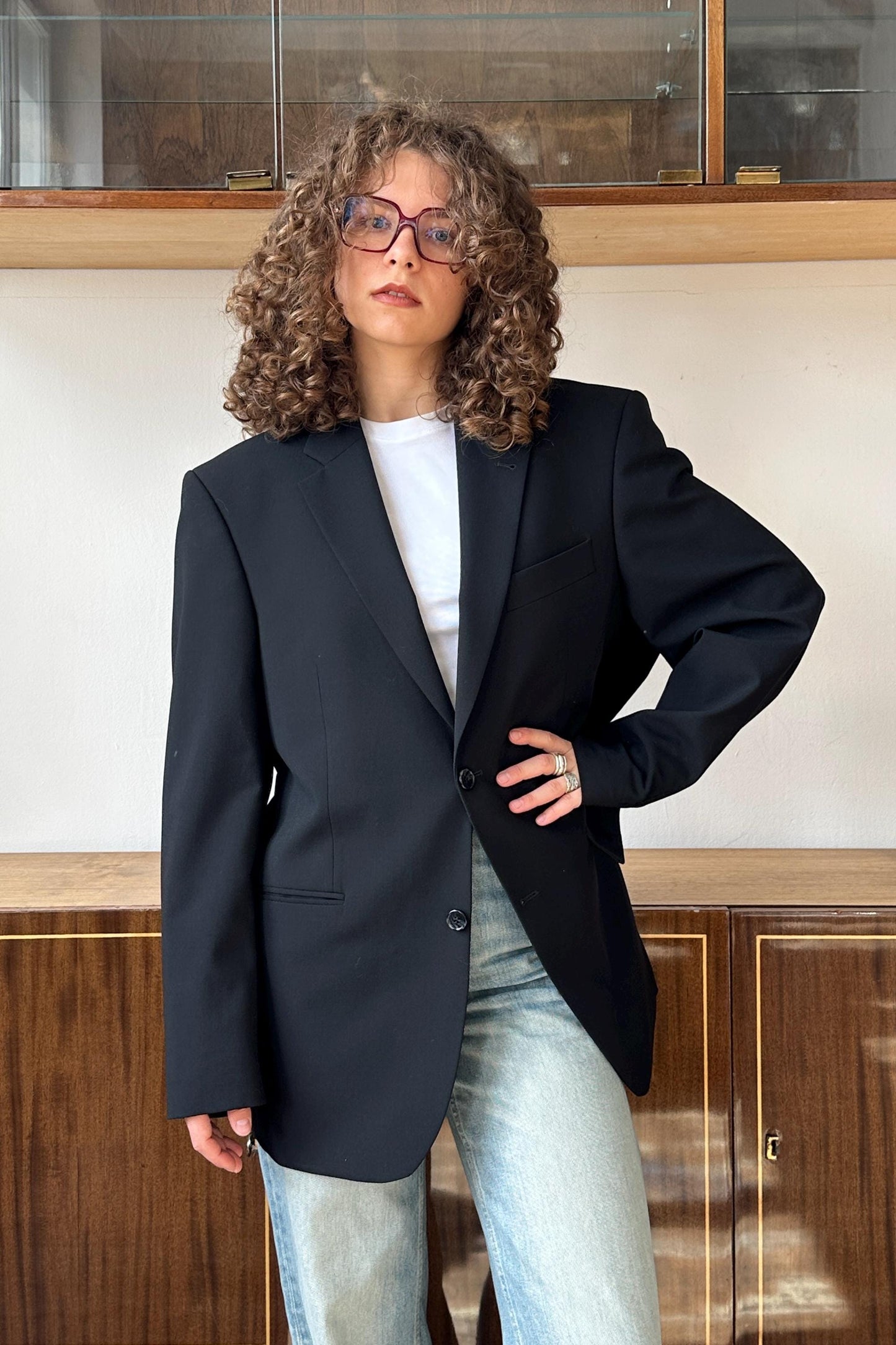 A woman with curly hair wearing a black blazer and jeans stands in front of a wooden cabinet.