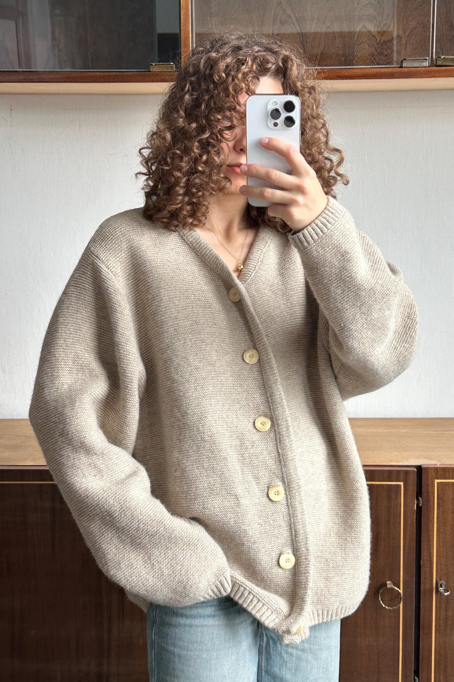 A person with curly hair is taking a selfie with a smartphone, wearing a beige cardigan and blue jeans.