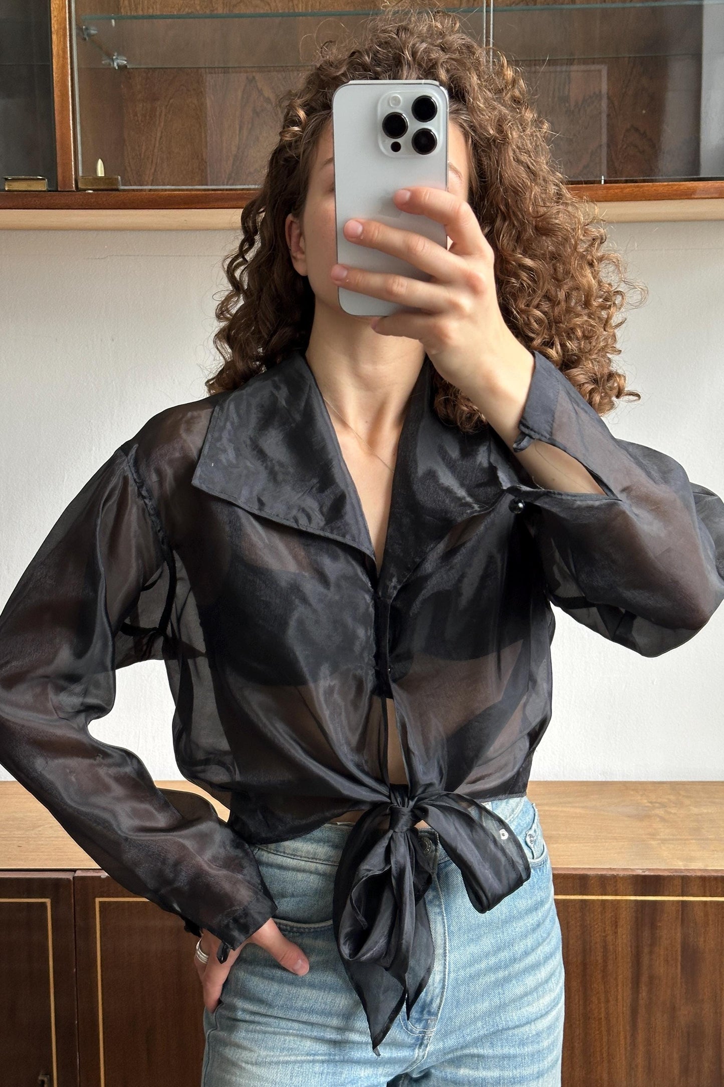 A woman with curly hair is taking a selfie with a smartphone, wearing a black blouse and blue jeans.