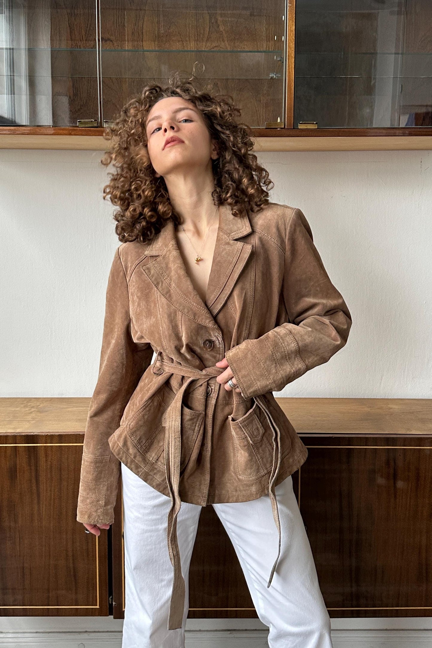 A woman with curly hair wearing a tan jacket and white pants stands in front of a wooden cabinet.