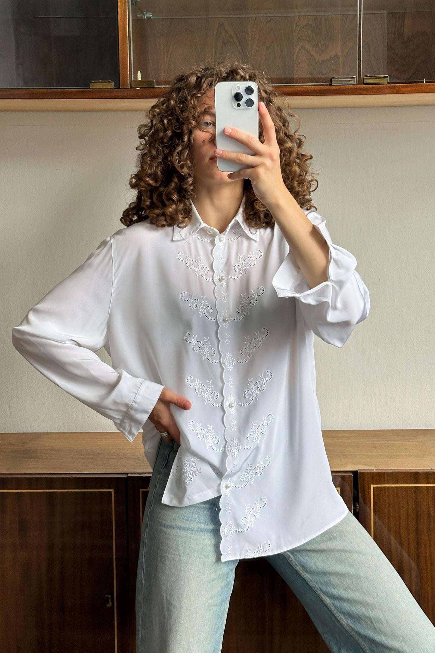 A person with curly hair is taking a selfie with a white iPhone, wearing a white blouse with embroidered details and light blue jeans.