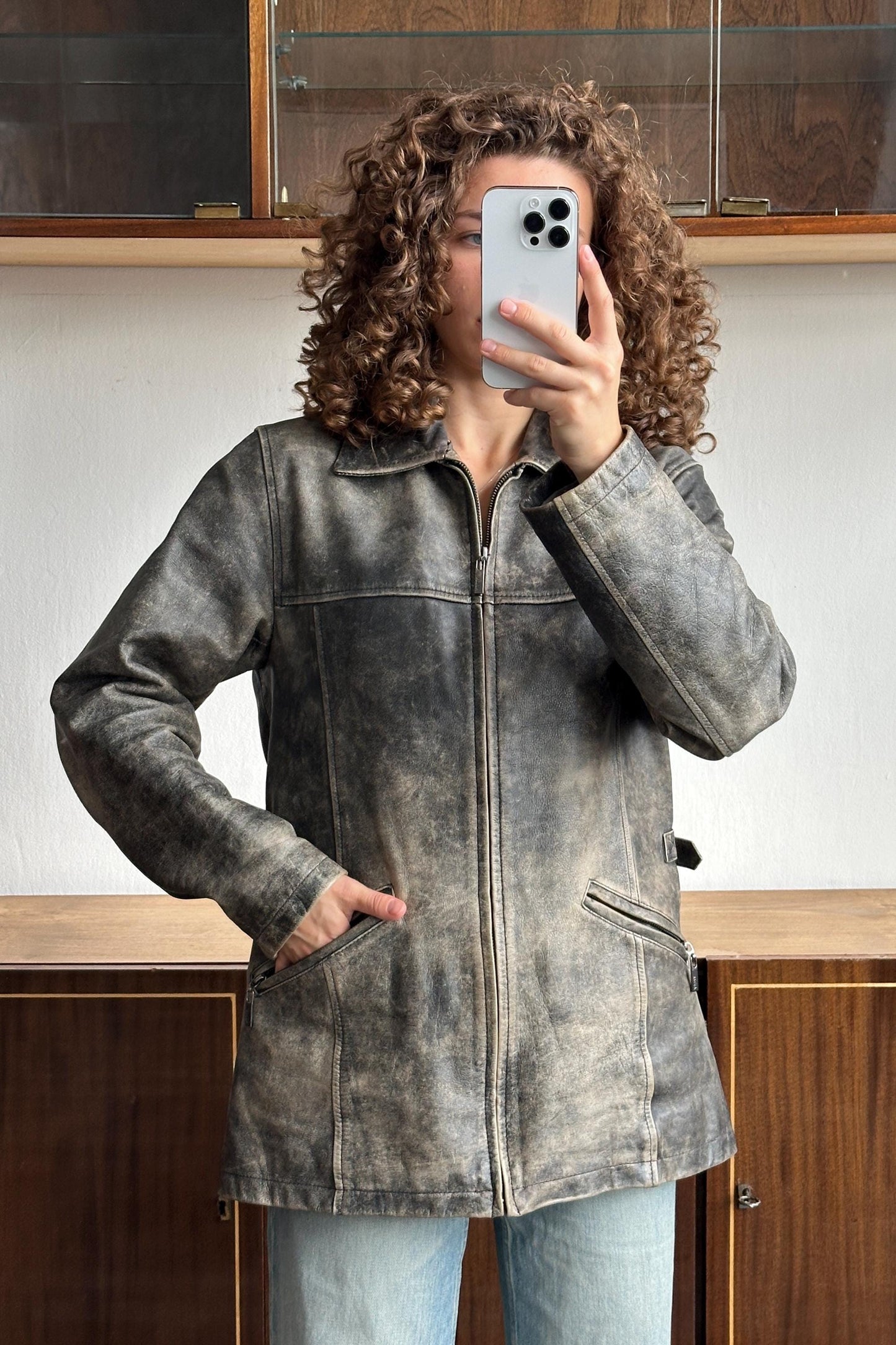 A person with curly hair is taking a selfie with a smartphone, wearing a gray leather jacket.